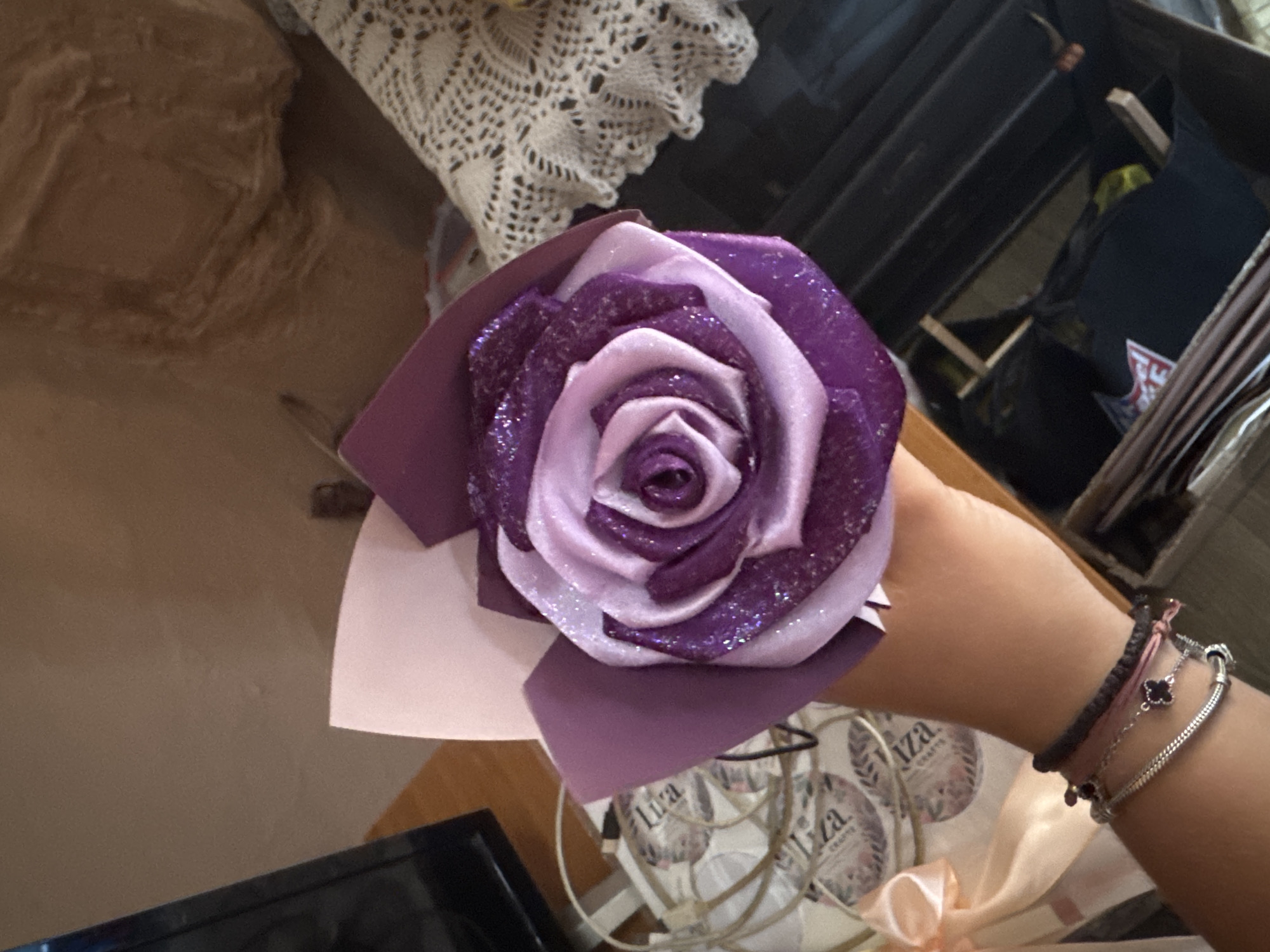A close-up shot shows a person's hand holding a handmade rose made of purple and pink ribbon with glitter. The rose is partially wrapped in purple and pink paper. In the background, a white lace tablecloth is visible, along with a partial view of a television and a woven basket containing various items, including cords and what appear to be labels with the word "Liza". The scene is set indoors, with a wooden floor and a wall in the background. There is no information in the image to indicate the location in the city of Sredets, Bulgaria.