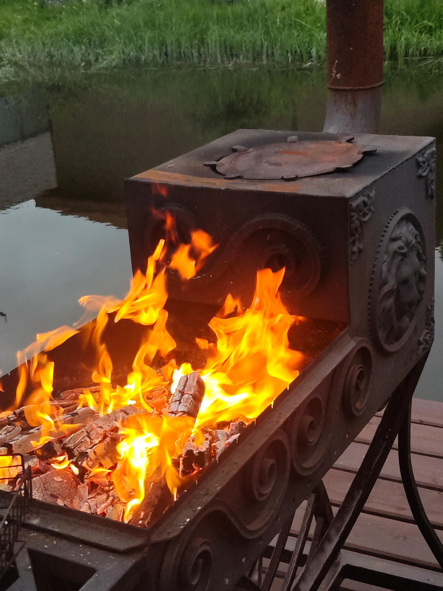 A square, black, metal grill is positioned outdoors on a wooden deck. The grill is filled with burning wood, producing bright yellow flames. The grill's design includes decorative circular and spiral patterns, along with a lion's head embossed on one side. A rusted metal chimney rises from the top of the grill. Behind the grill, there is a body of water reflecting the surrounding landscape, including green grass. The location is in Chaikovsky, Russia.