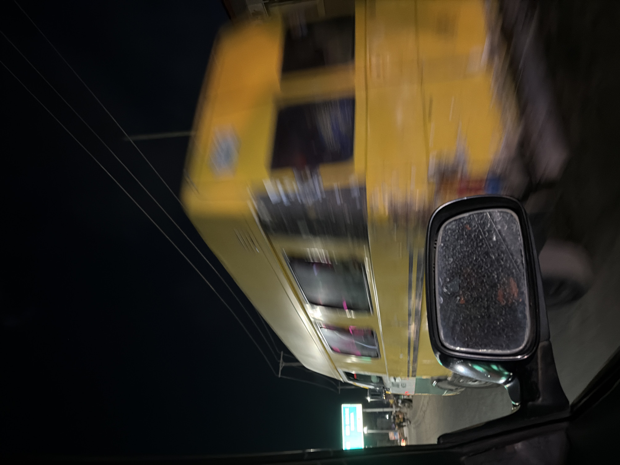 A yellow bus is speeding alongside a car. A side mirror of the car is visible, with water droplets on its surface. In the background, street lights and a green sign are present, suggesting a commercial area at night. The location is possibly in Lagos, Nigeria, based on the bus type and environment.