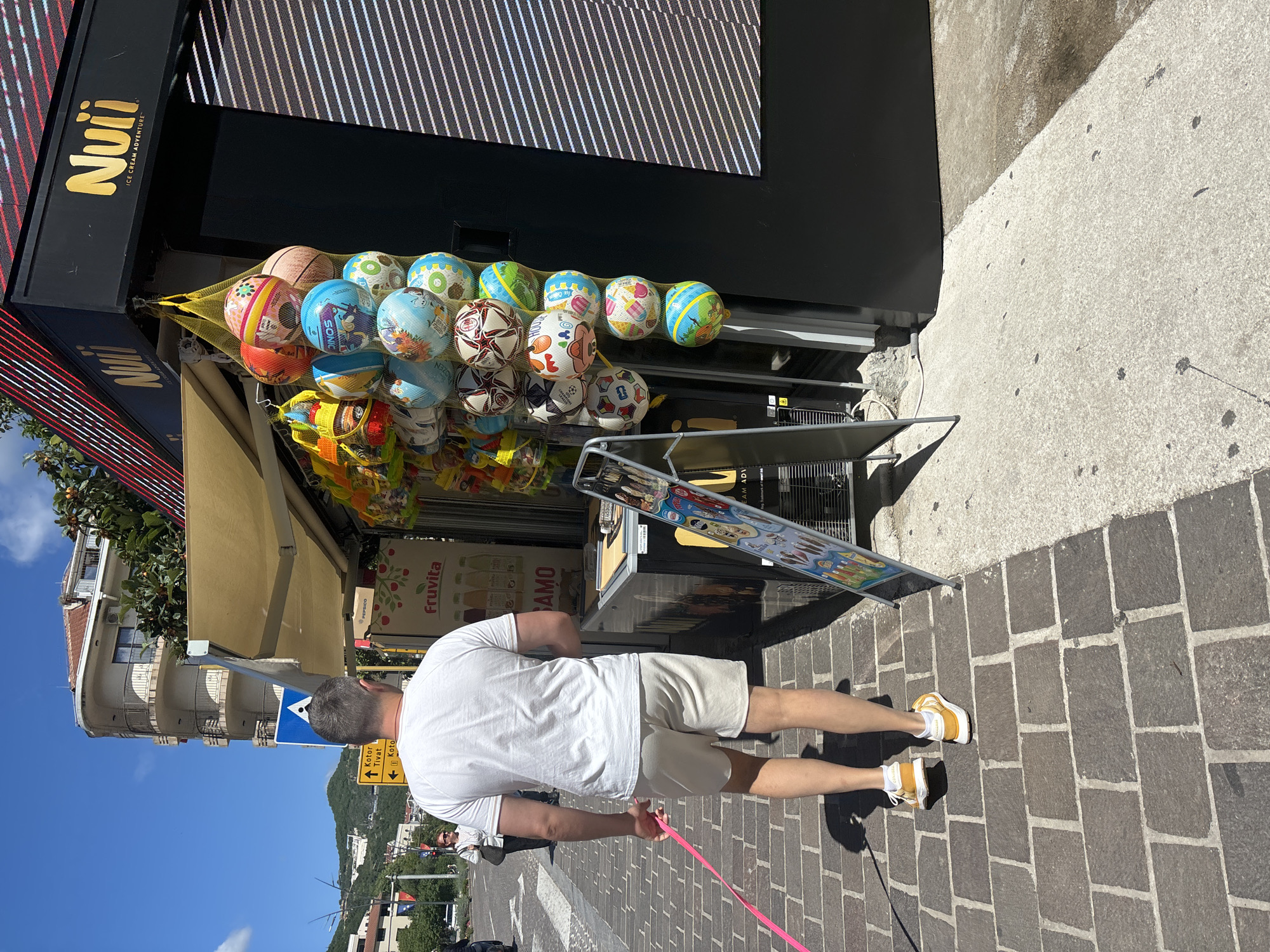 A man wearing a white t-shirt, tan shorts, and yellow and white sneakers stands on a cobblestone street, holding a pink dog leash. He is facing away from the camera, looking toward a shopfront. The shop, identified by a sign reading "Nuti ICE CREAM ADVENTURE", has a display of colorful inflatable balls in a net hanging above a small stand with tourist-oriented advertisements. A road sign indicating directions to Kotor and Tivat is visible in the background, along with buildings on a hillside, placing the scene in the city of Budva, Montenegro.