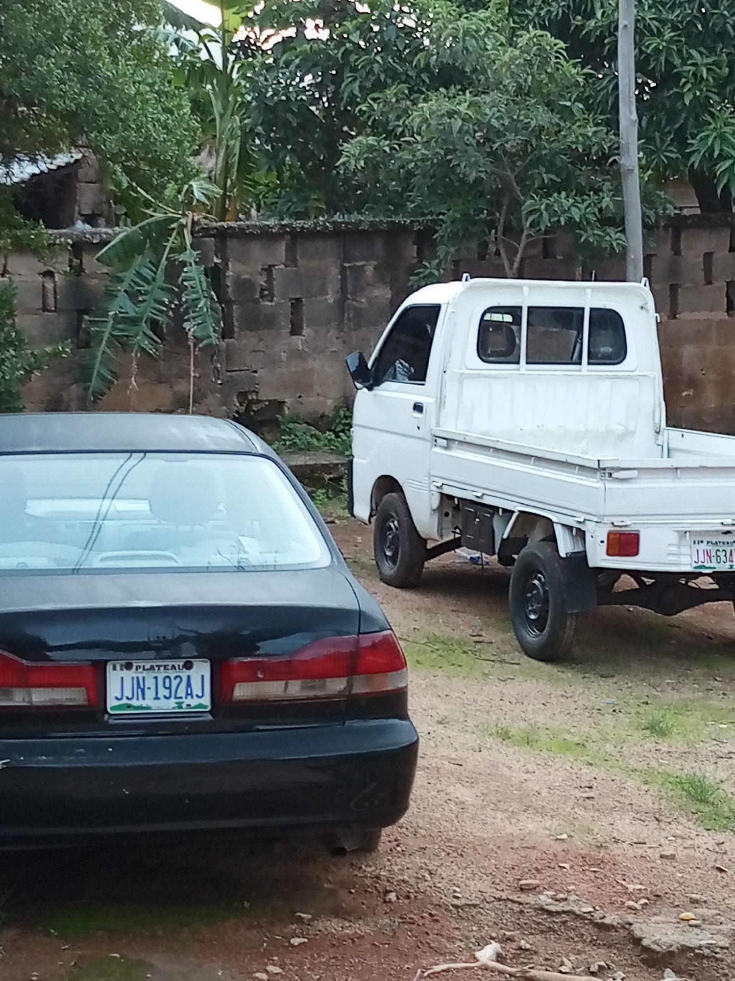 The image depicts an outdoor scene likely in Jos, Nigeria, with two vehicles parked on a dirt and gravel surface. In the foreground, the rear of a black sedan is visible, displaying a license plate that reads "JJN-192AJ". Behind it, a white pickup truck is parked, also showing a license plate reading "JJN-634...". The vehicles are situated in front of a low, brick wall, beyond which lush green trees and foliage are abundant. The overall environment suggests a residential or semi-rural setting. The lighting indicates it is daytime. No people are clearly visible in the frame.