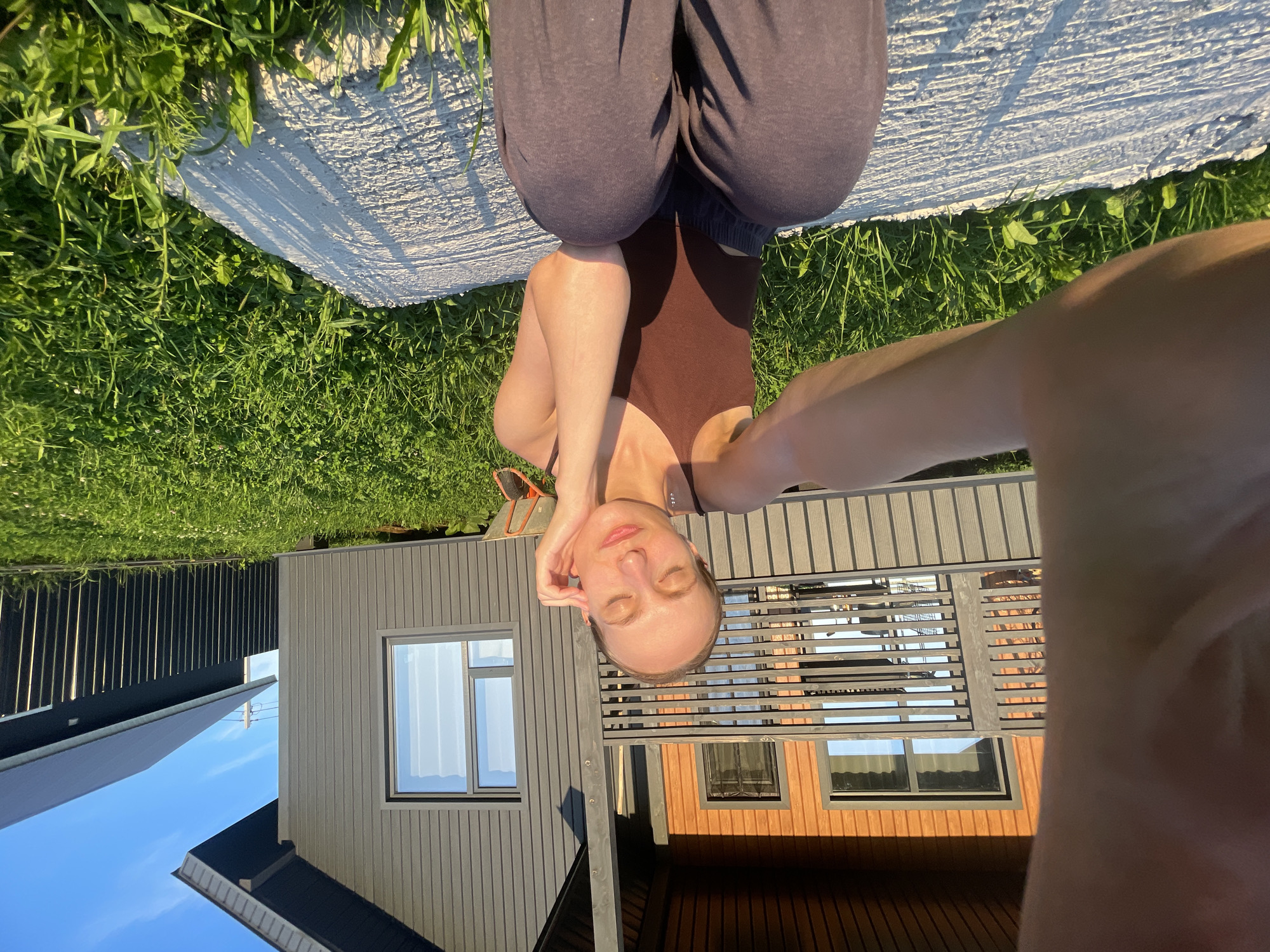A young woman with blonde hair tied back is sitting on a concrete slab in the grass, facing the camera and taking a selfie. She is wearing a brown tank top and dark grey sweatpants. Her eyes are closed and she has her right hand touching her cheek, with her left arm extended towards the camera. Behind her is a wooden house with brown siding and a grey exterior. A green lawn with scattered white flowers is visible around the concrete slab. In the background, there is a black fence and another dark-colored building. The sky is clear and blue, with the sun casting shadows on the grass and concrete. The scene appears to be in a rural or suburban area, likely in the village of Podshivalovo, Russia, given the architectural style and the presence of a dacha-like structure.