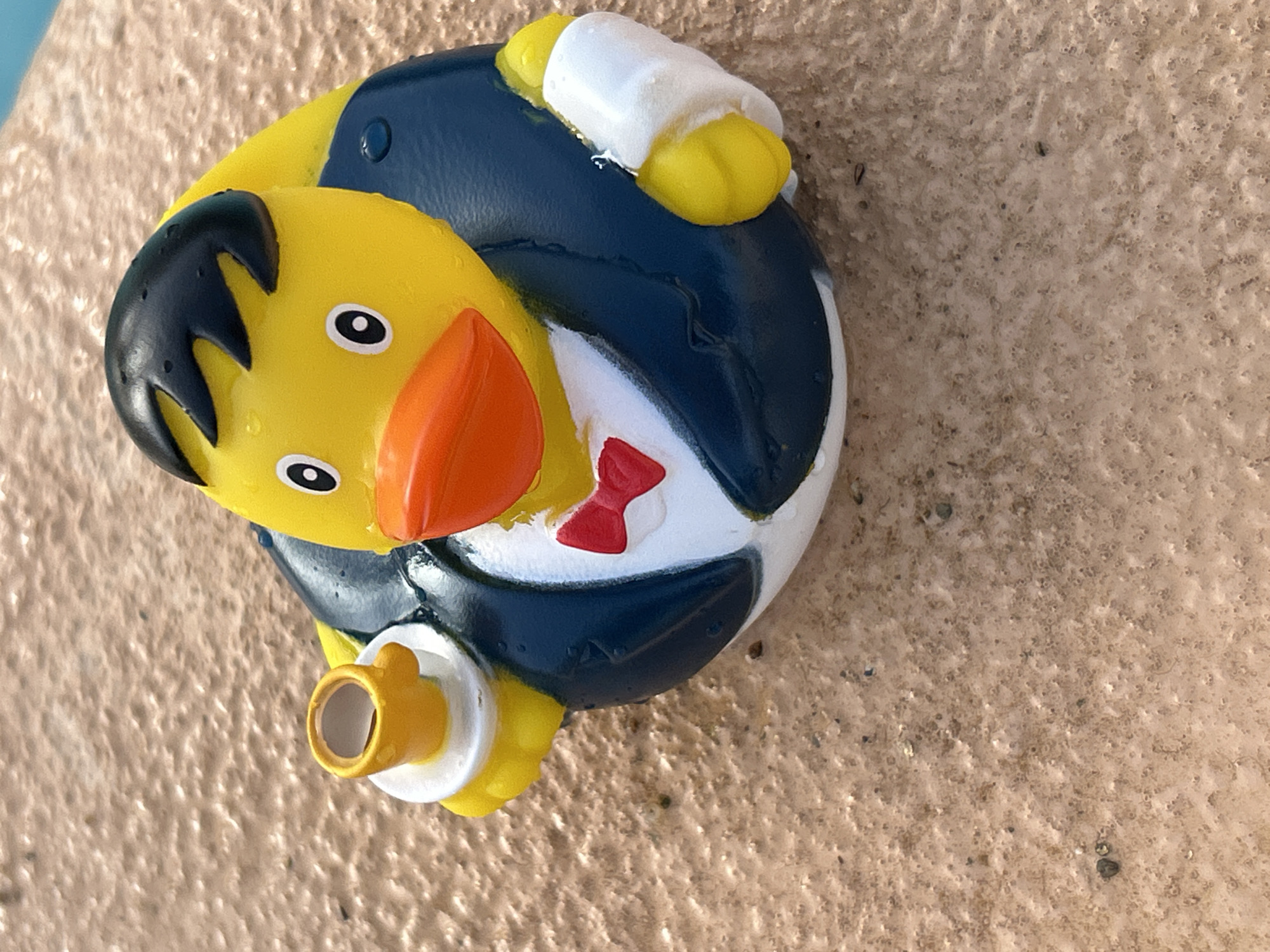 A rubber duck toy, designed to resemble a duck wearing a suit and holding a cup of coffee, rests on a textured, sandy surface. The duck is primarily yellow with a black hairstyle, a red bow tie, and a navy blue suit. Its wings are depicted as yellow with white cuffs. The sandy surface appears to be outdoors, possibly near a body of water given the presence of what looks like a blue surface in the upper right corner of the image. The lighting suggests it is daytime. The exact location in Sant Vicenç de Montalt, Spain, cannot be determined from the image alone.