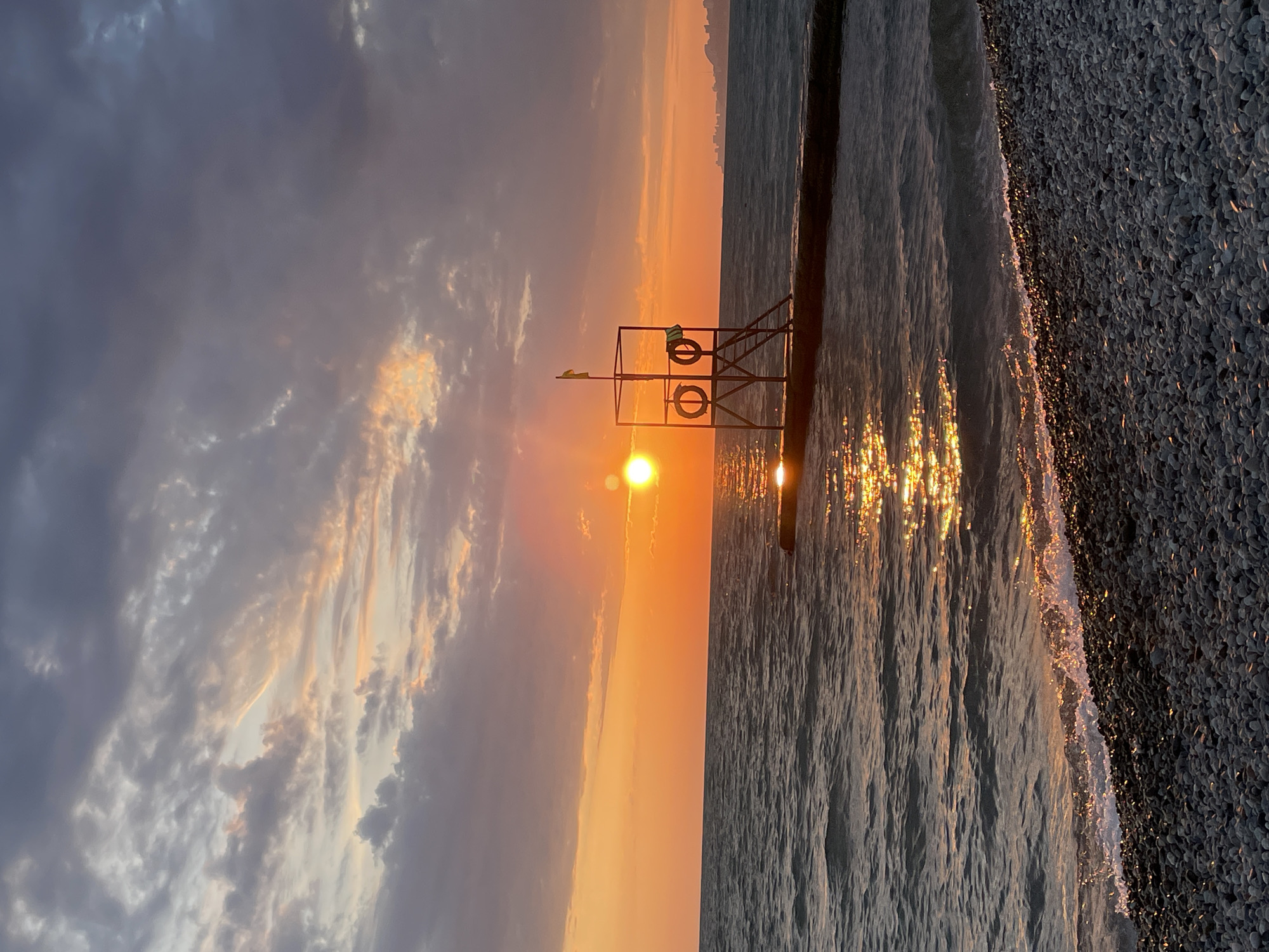 A vertical, eye-level, medium shot captures a sunset over the sea in Sochi, Russia. The sun, a bright orange orb, is low in the sky, casting a golden reflection on the rippling water. In the foreground, a pebble beach meets the gentle waves. A concrete pier extends into the sea, supporting a lifeguard stand with two life preservers and a yellow flag. The sky is filled with a mix of grey and pink clouds, tinged with the warm hues of the setting sun. In the distance, a city skyline is faintly visible on the horizon. The scene is devoid of any people, creating a serene and tranquil atmosphere.