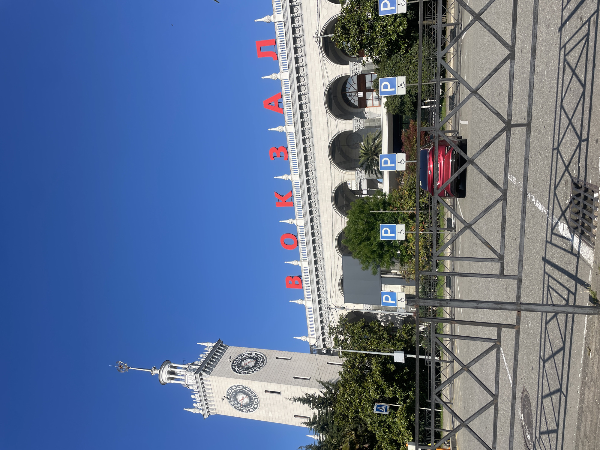 The image depicts the Sochi Railway Station in Sochi, Russia, on a clear, sunny day. The station's tower clock is visible, as is the façade with the red Cyrillic letters spelling out "ВОКЗАЛ," the Russian word for train station. Parking signs are present, and a red car is parked near the station. A dark metal fence is in the foreground, casting shadows on the asphalt ground. The environment consists of trees around the building and parking areas.