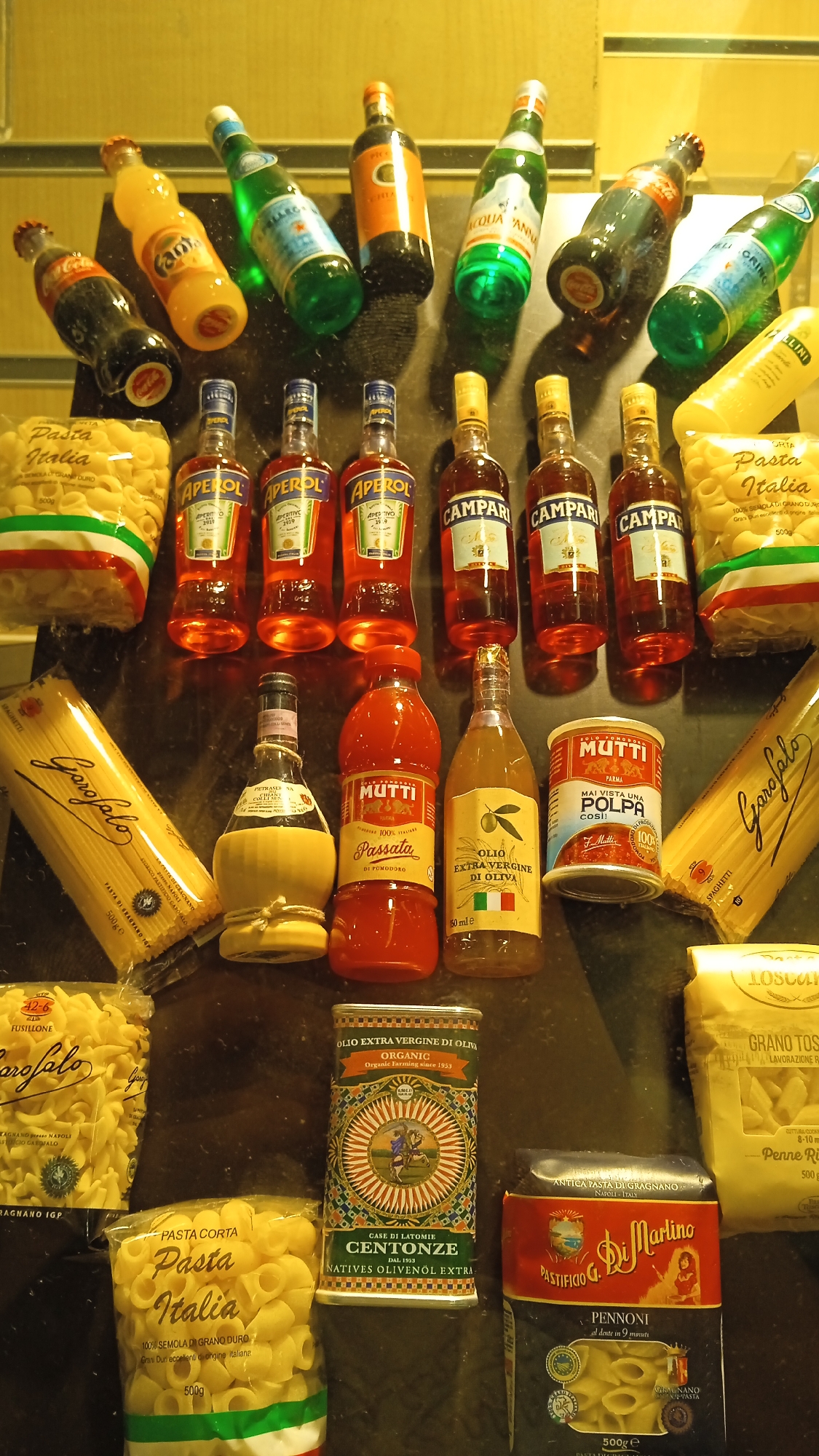 The image shows an overhead view of a collection of Italian food and beverage products displayed on a dark, reflective surface. The items include several bottles of Aperol and Campari, as well as bottles of Fanta, Coca-Cola, San Pellegrino, and Acqua Panna. There are also packages of pasta, jars of passata and olive oil, and a can of tomatoes. The products are arranged in rows and clusters, with some items overlapping. The lighting in the scene is warm and casts shadows. The overall impression is that of a curated display of Italian culinary items. There is no discernible location in Milan or any other city, nor any people or actions depicted.