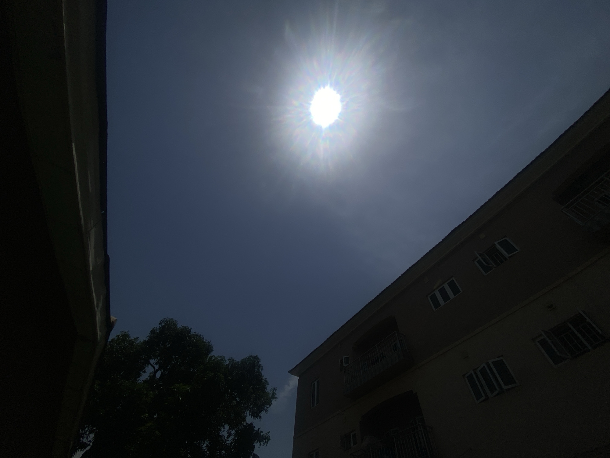 The image depicts a daytime scene in Barden Yana, Nigeria. The sun is high in the sky, appearing as a bright, spherical light source. To the left is a portion of a building, possibly a residential structure. In the lower left, a tree with dense foliage. To the right, a three-story residential building is visible, with white-framed windows and balconies. The sky appears dark blue. No people are visible in the frame.