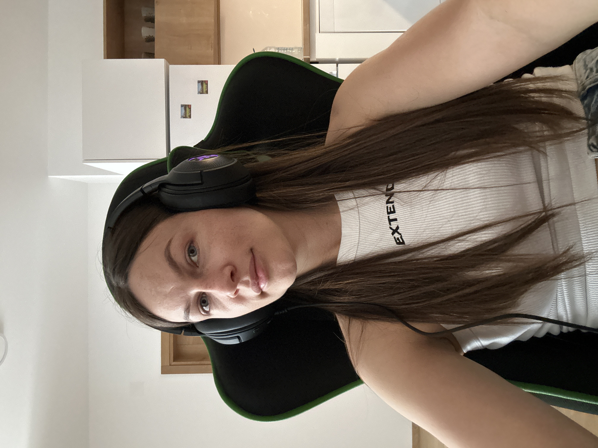 A woman sits in a black and green gaming chair wearing a white tank top with "EXTEND" printed on it and black headphones. She has long brown hair and is looking at the camera. The background includes white kitchen cabinets, a wooden cabinet, and a wall. The image does not contain enough information to determine the location in Budva, Montenegro.