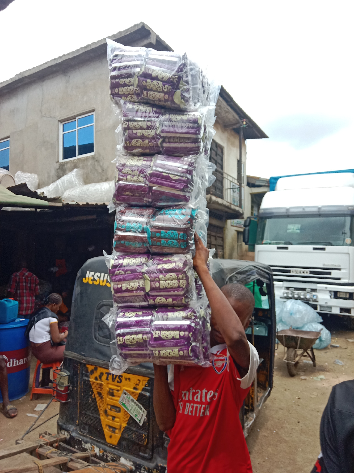 In Onitsha, Nigeria, a man wearing a red Arsenal jersey carries a tall stack of packaged goods on his head. The goods are stacked high and wrapped in clear plastic. He stands next to a black and yellow TVS tricycle. A white Iveco truck is parked in the background. Several people are visible in the background, including a woman sitting on a red stool near a blue barrel. The environment suggests a busy market or commercial area.