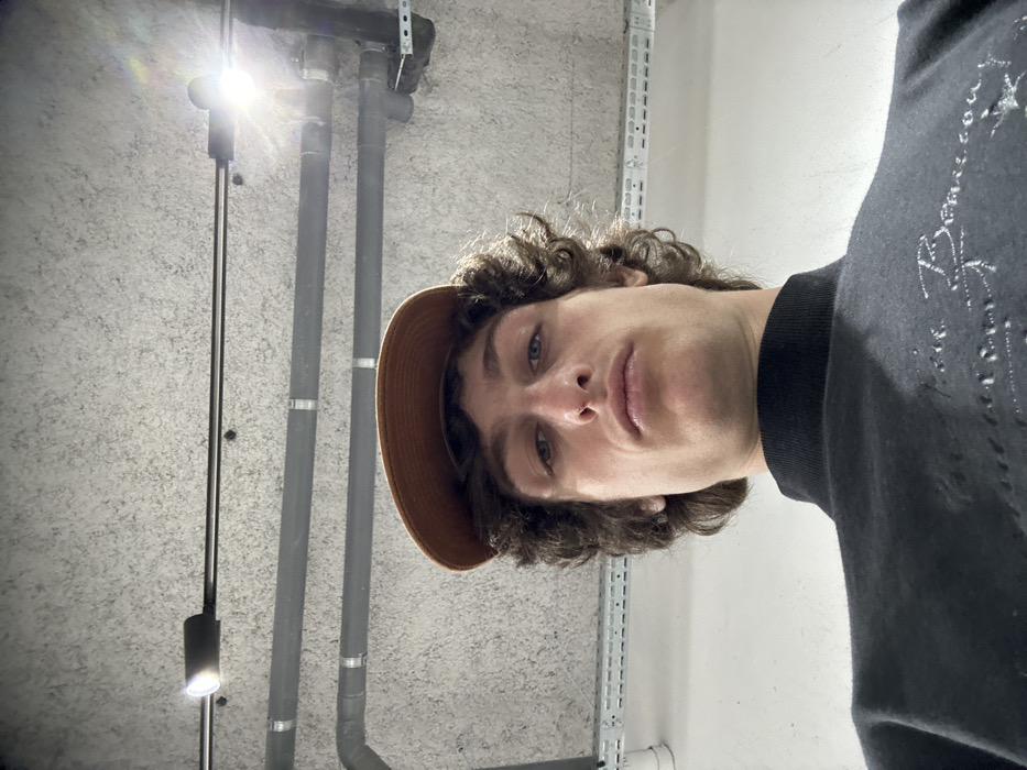 The image displays a male individual photographed from a low-angle perspective, looking directly upwards into the camera. He has brown, curly hair and wears a brown baseball cap with the brim facing forward. He is dressed in a black high-neck long-sleeved top featuring light-colored embroidery or print on the chest. His expression is neutral. The background consists of a textured, light grey concrete ceiling. Exposed utility infrastructure is visible, including multiple dark grey pipes secured by metal clamps and brackets, running horizontally across the upper portion of the frame. A horizontal metal track or conduit is also present, equipped with at least two bright light fixtures, one of which on the right exhibits significant lens flare. The environment indicates an indoor, utilitarian setting, consistent with a parking garage, basement, or service area. The specific location within Joinville-le-Pont, France, is not identifiable from the visual content; however, the scene details suggest an urban infrastructure environment.