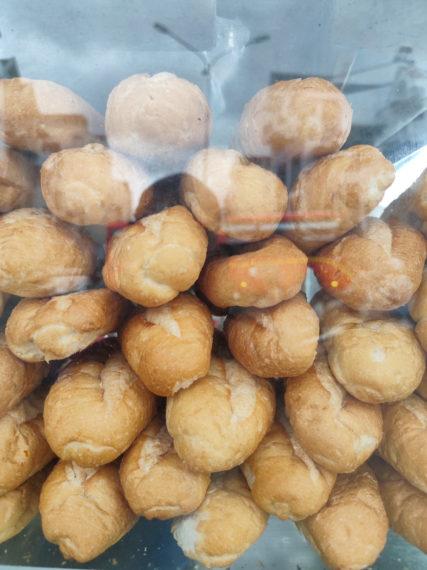 A stack of small, golden-brown bread rolls is displayed behind a glass pane. The rolls are plump and have a slightly textured surface, with some showing a visible split or crease down the center. They are piled haphazardly on top of each other, filling the frame from edge to edge. The lighting is bright, casting a soft glow on the bread and highlighting its warm color. Reflections on the glass suggest an indoor environment, possibly a bakery or a market stall in Da Lat, Vietnam, a city known for its French colonial architecture and vibrant food scene. The scene is devoid of people, focusing solely on the appetizing presentation of the baked goods.