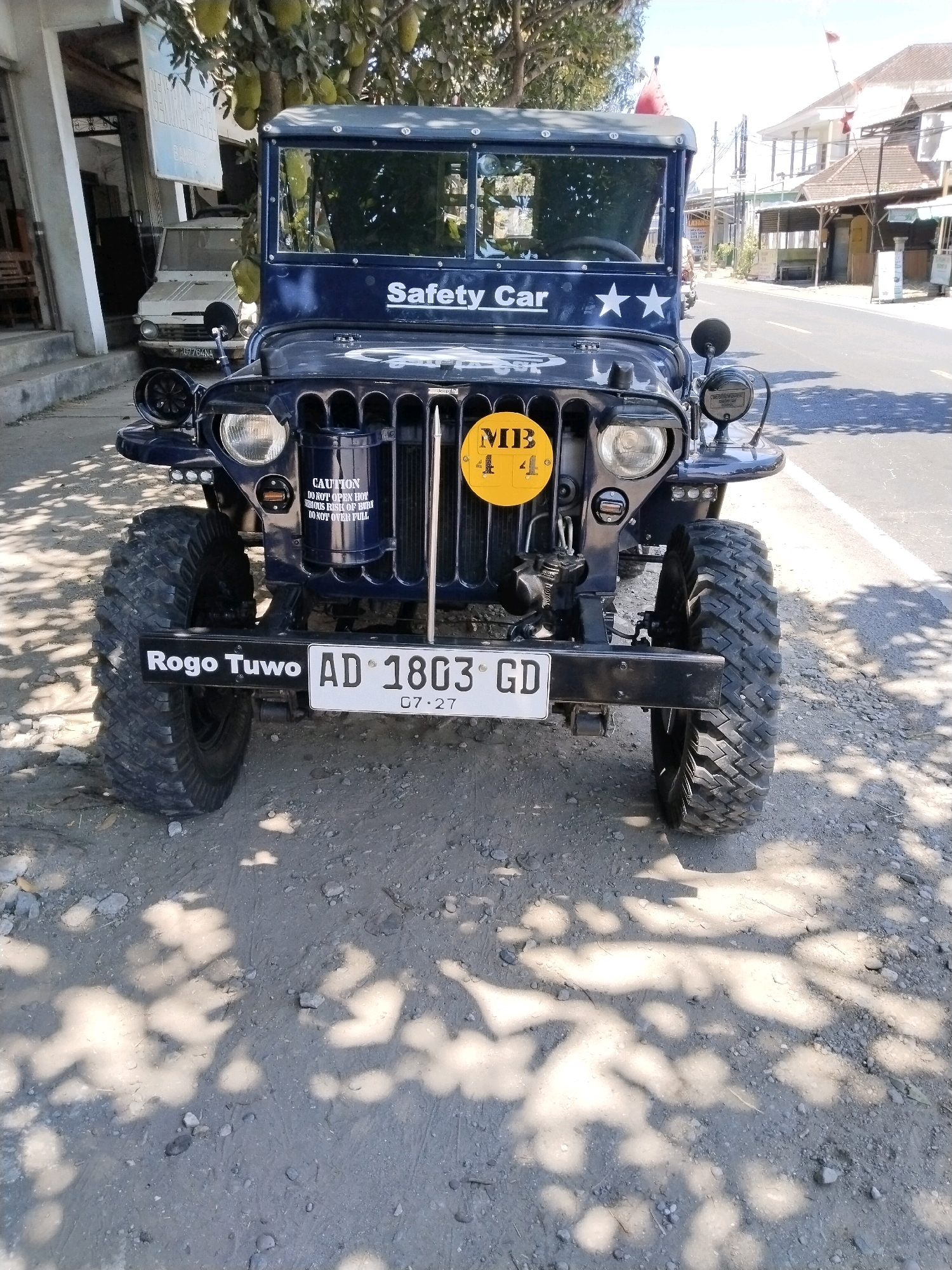 A vintage dark blue Jeep, labeled "Safety Car," is parked on a dirt road in front of a building in Mergayu, Indonesia. The Jeep has a yellow circular emblem on its front, reading "MB 4 4." A white license plate on the front bumper reads "AD 1803 GD" and "07.27." The vehicle's headlights and turn signals are visible. A white van is parked behind the Jeep to the left. The scene is illuminated by sunlight, casting shadows from trees onto the road. Buildings and a street are visible in the background.