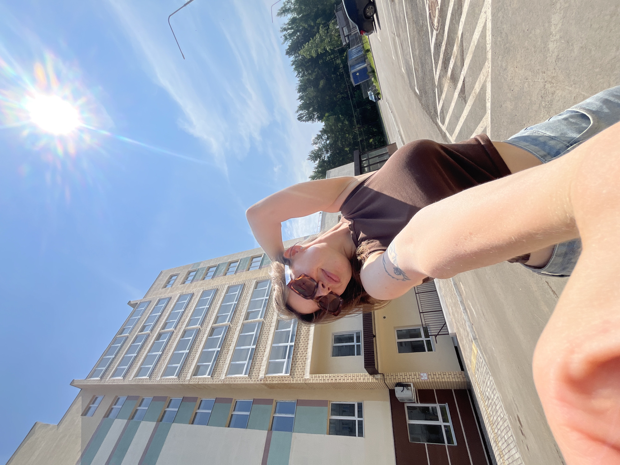 An outdoor, low-angle, full-body, close-up selfie is taken on a sunny day in the city of Chaikovsky, Russia. The background features a modern apartment building with beige brick and cream-colored facade, and green and brown decorative stripes. The building has multiple floors with numerous windows. The sky is clear blue with a bright sun causing lens flare and a visible rainbow halo. A portion of a person's arm and hand are in the foreground, obscuring the camera lens. The person in the selfie, viewed from the chest up, is wearing a brown tank top and light blue jeans, and dark sunglasses. They have their right arm raised, hand behind their head, and are looking at the camera. Behind the apartment building, a line of green trees is visible, and beyond that, a parking lot with a black car, a dark blue utility structure, and a street light. The ground in the foreground is asphalt with painted parking lines and a yellow pedestrian ramp. The lower portion of the apartment building has an air conditioning unit mounted on the wall.