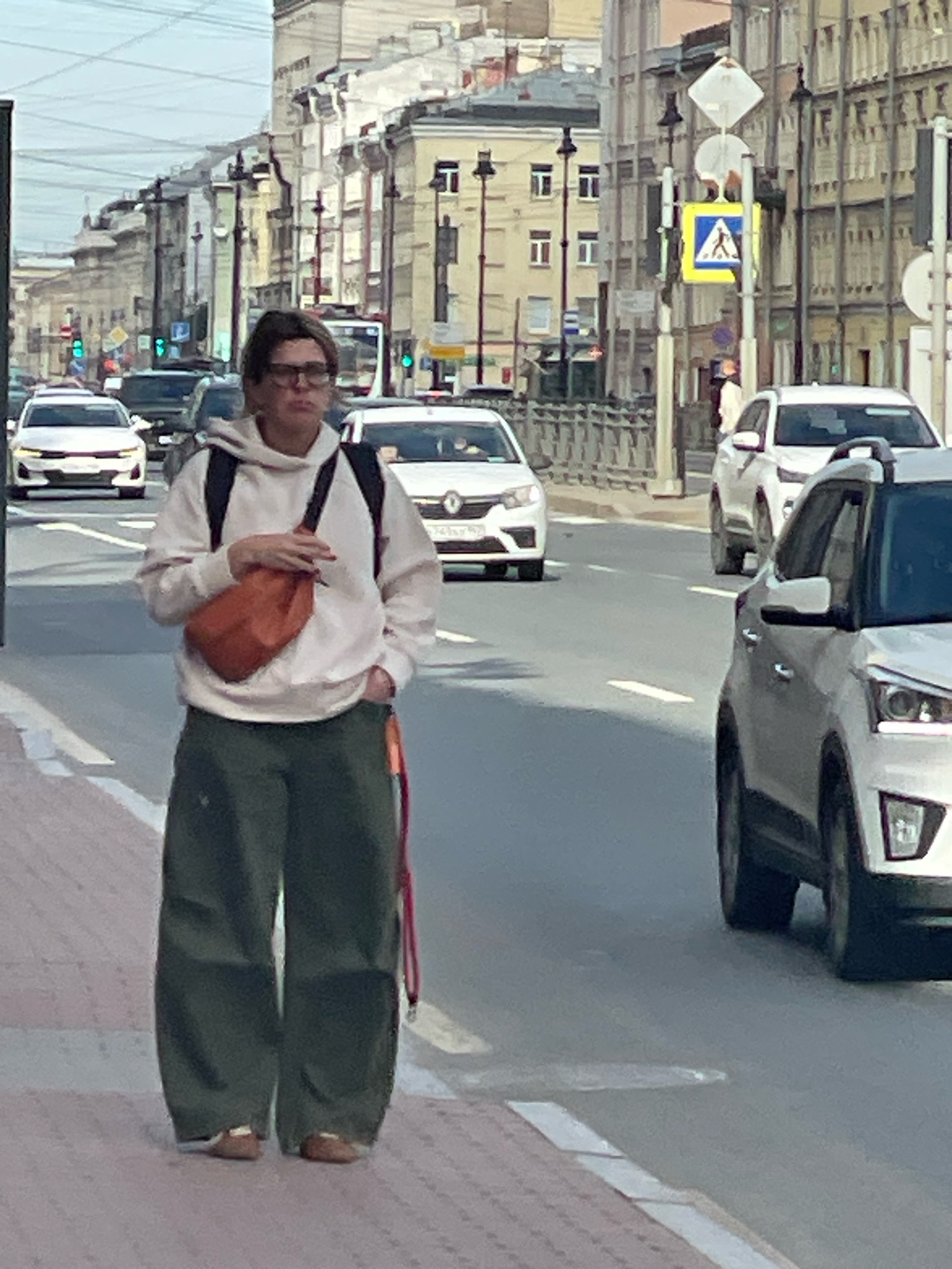 The image depicts a street scene in Saint Petersburg, Russia, with a person in the foreground walking along a sidewalk. The person, who appears to be wearing glasses and a light-colored hooded sweatshirt, is carrying an orange bag across their chest and has dark green pants. They are walking on a sidewalk next to a street with several cars. In the background, there are buildings lining the street, a traffic light, and street signs, including a pedestrian crossing sign. The sky is overcast.