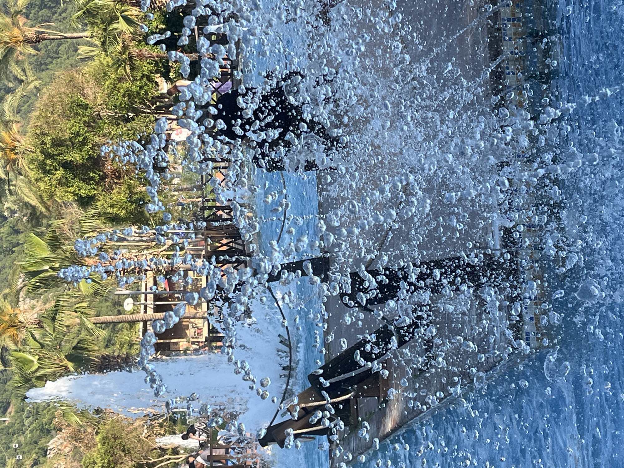The image shows a water fountain in Alanya, Turkey. The fountain is shooting water high into the air, with many individual droplets visible. In the background, palm trees and other greenery can be seen, along with a glimpse of buildings and people. The fountain is located near a tiled basin. People are visible on a walkway next to the fountain.