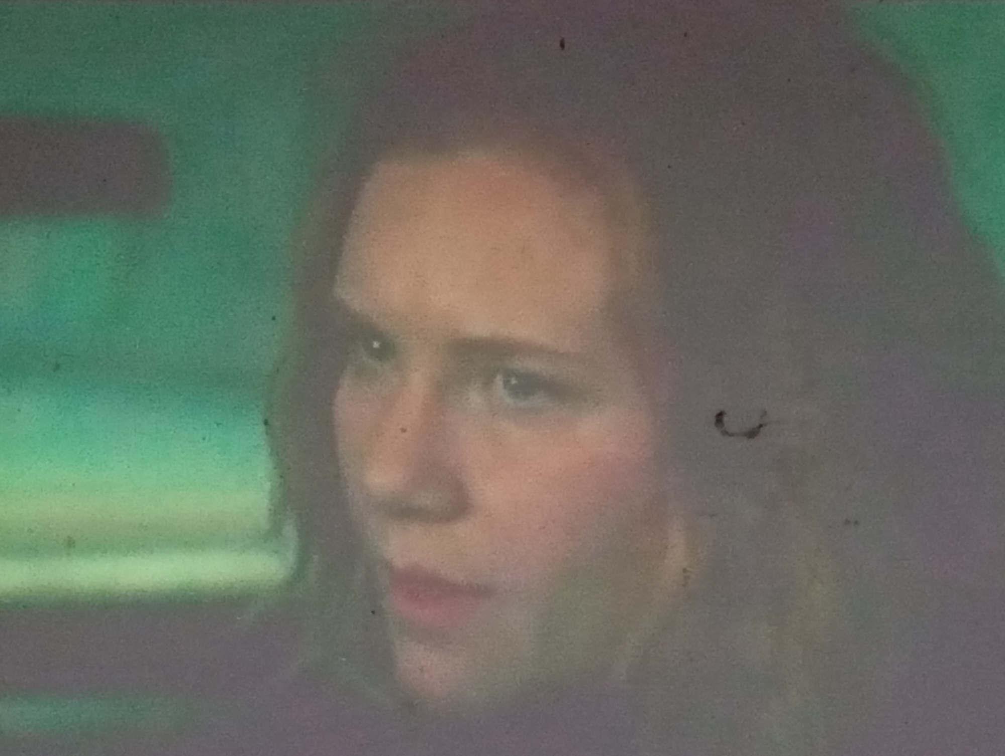 This is a close-up, grainy, medium-shot of a young woman's face, partially obscured by a greenish-gray tint. She appears to be looking slightly to her left, with her mouth slightly open. Her skin is fair, with rosy cheeks. Her hair is dark and appears to be pulled back or is naturally curly. The background is blurred and predominantly green, with some darker, indistinct shapes. There are a few small dark specks visible on the image, suggesting a poor quality recording or the presence of dust. The overall lighting is dim. There is no discernible information within the image to indicate its location in Sofiivska Borshchahivka, Ukraine.