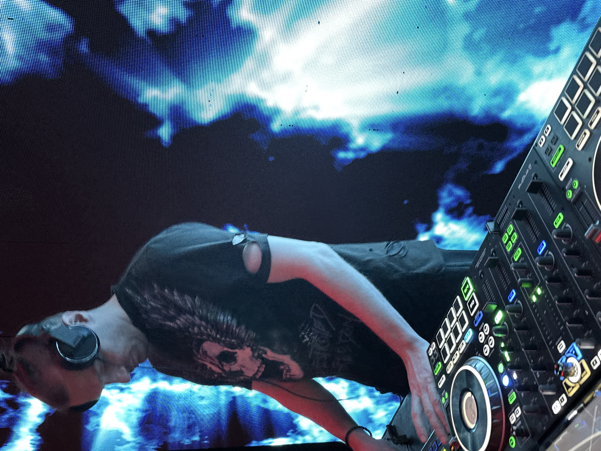 A person with headphones is operating a DJ controller in front of a large display screen. The person is wearing a dark t-shirt with a graphic on it. The screen shows a blue and white abstract design, potentially a light show or visual effect. The controller has multiple buttons, knobs, and sliders, with some of the controls illuminated. The location is unknown; information about Votkinsk, Russia is not visible in the image.