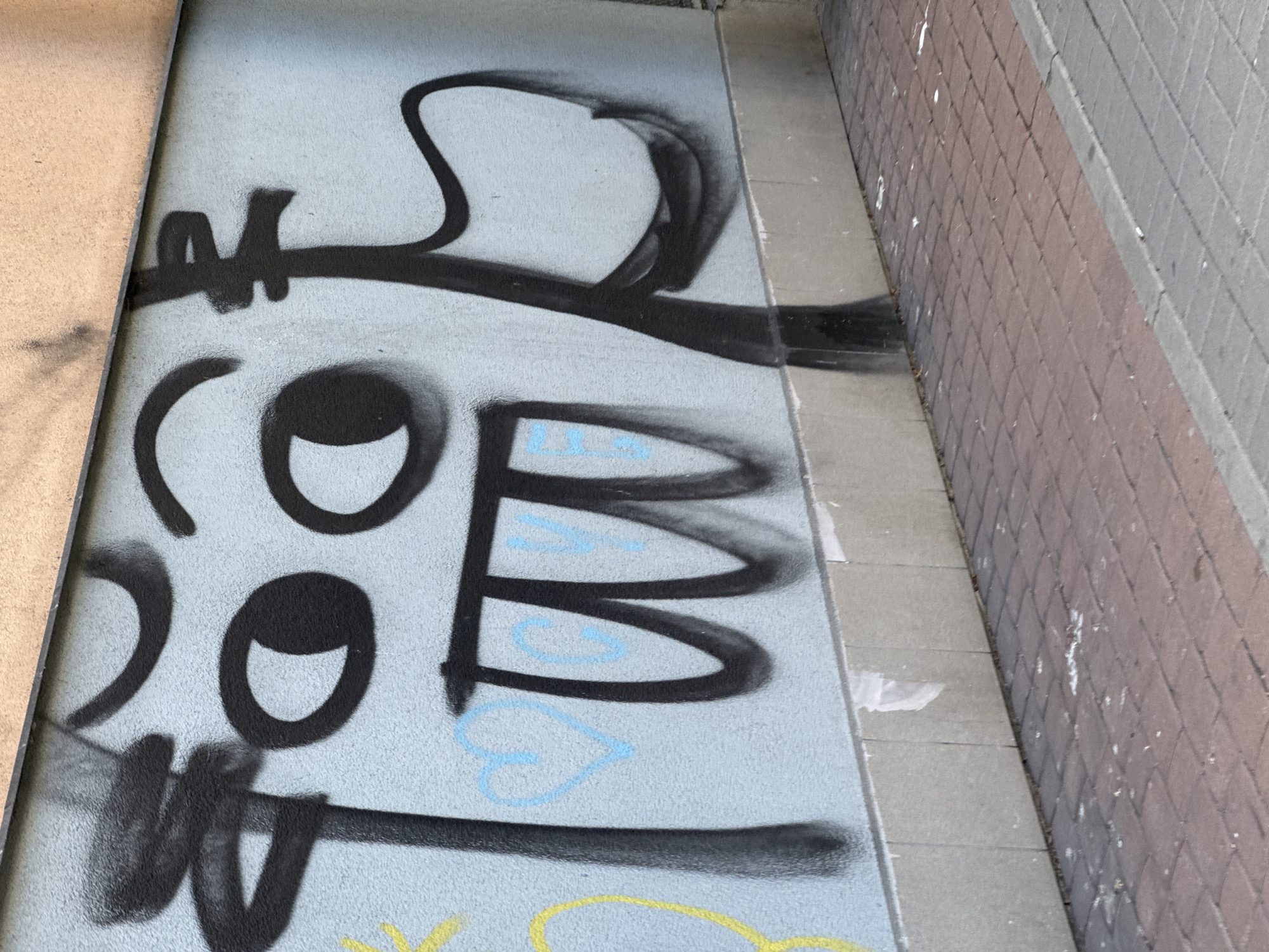 The image shows a section of a light blue concrete wall with black graffiti. The graffiti is stylized and appears to depict a cartoonish face with large eyes and a wide, open mouth. There are also some light blue spray-painted details on the wall, including what appears to be a heart and some letters. The wall is at the base of a building with an orange facade visible at the top. The ground in front of the wall is paved with red brick pavers. The scene is outdoors, possibly on a street or walkway. No people are visible in the image.