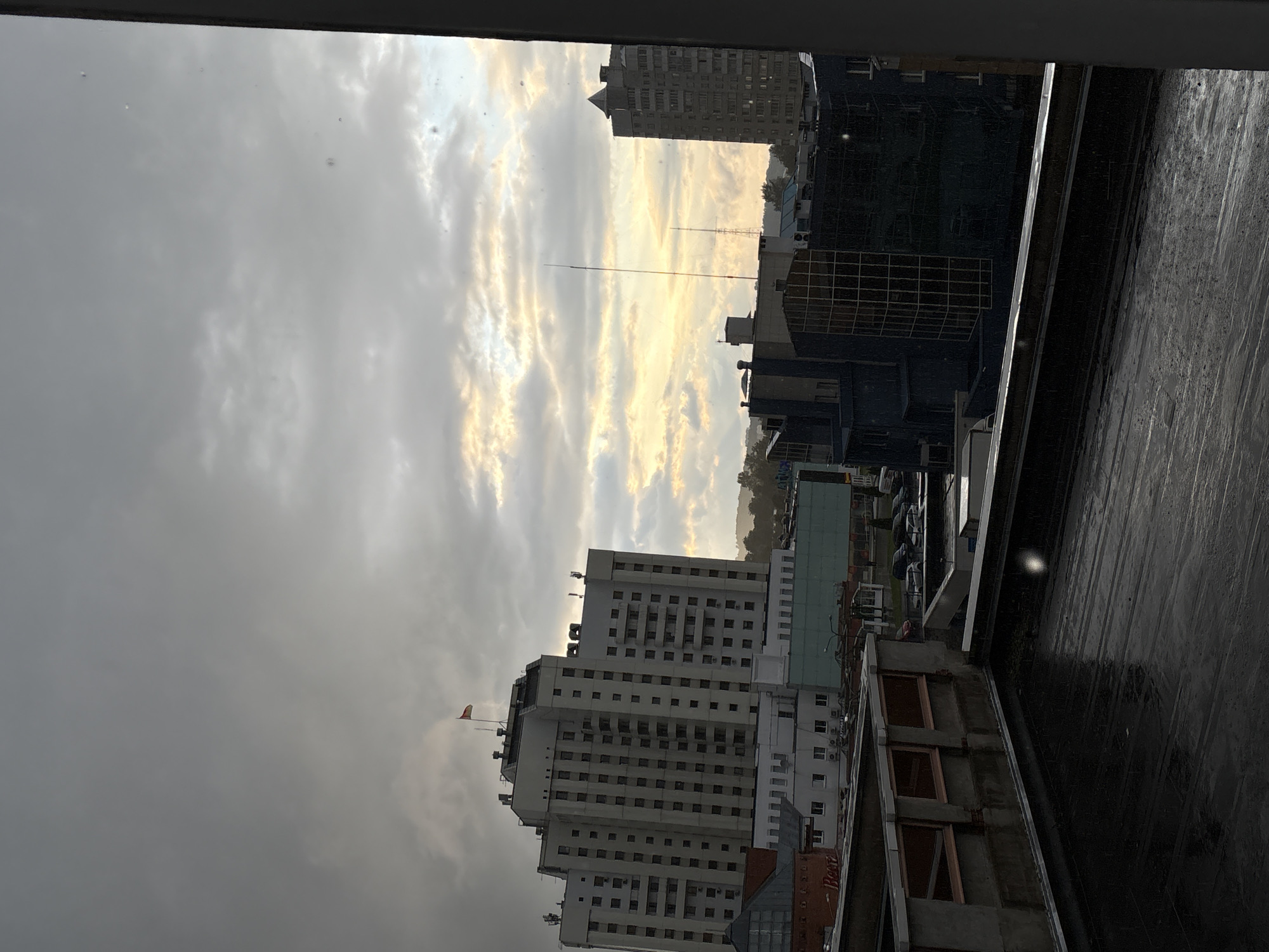 The image depicts an overcast sky over a city skyline, possibly Vitsebsk, Belarus, with several buildings visible. In the foreground, a dark, wet rooftop stretches across the bottom of the frame. Behind it, a low-rise building with glass windows and concrete structures can be seen. Further back, a tall, white, multi-story building dominates the left side of the image, with a flag atop its roof. To the right, other buildings of varying heights and architectural styles, including a darker, more modern building with a glass facade, are present. Two tall antenna-like structures are also visible in the distance. The overall impression is a slightly gloomy urban landscape under a cloudy sky. There are no people or specific actions visible in the image.