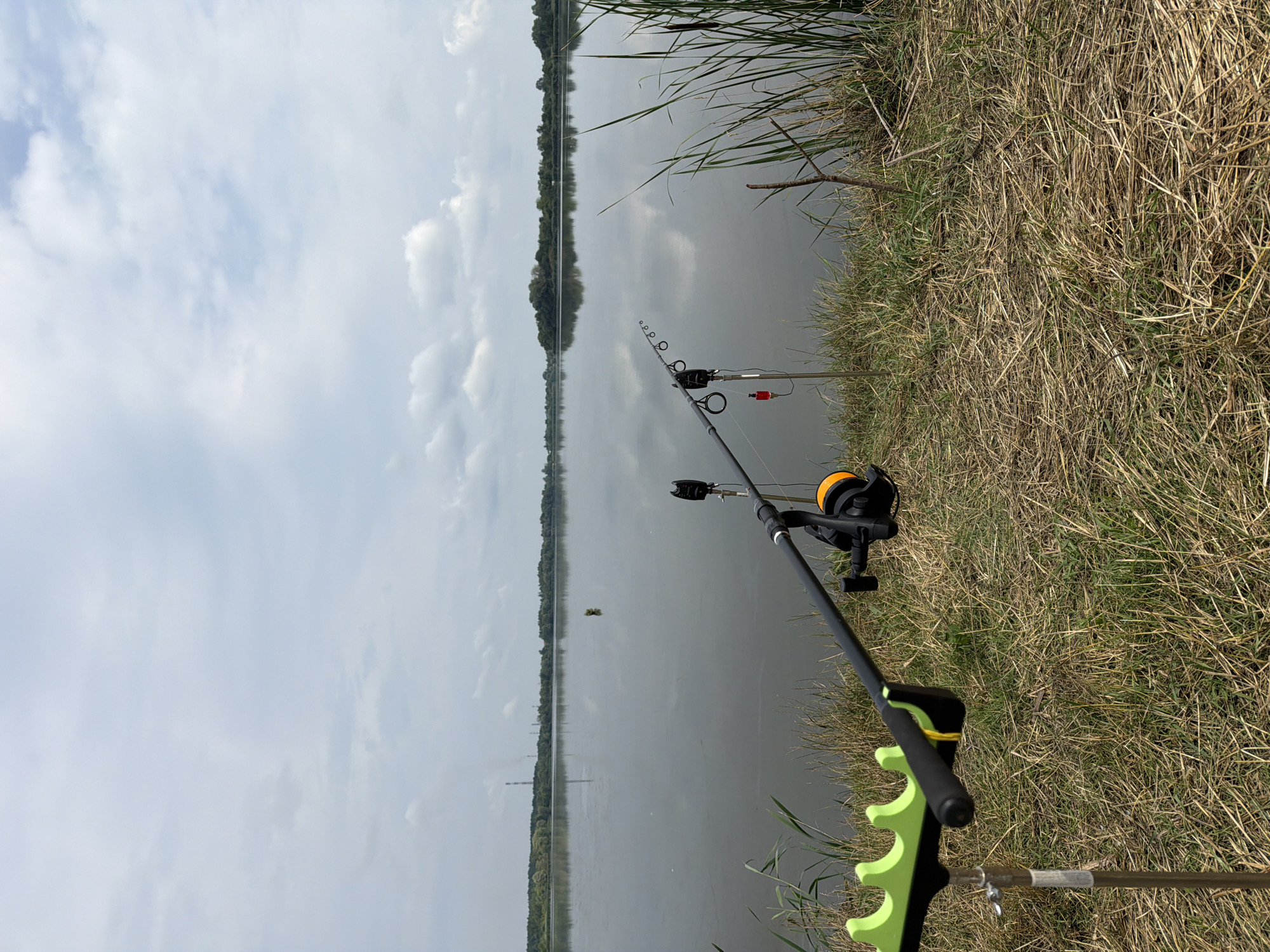 The image depicts a serene fishing scene by a large body of water. In the foreground, a fishing rod is set up on a stand, its reel visible. The rod rests on a green fishing rod rest, and a small red float hangs from the line. The ground is covered in dry grass and reeds, suggesting a rural or natural setting. The calm water reflects the overcast sky, which is filled with scattered clouds. In the background, a line of trees forms the horizon, and a distant industrial structure with smokestacks is faintly visible. The overall impression is one of quiet solitude and anticipation. There are no people visible in the image. The location is identified as the city of Ruda, Ukraine.