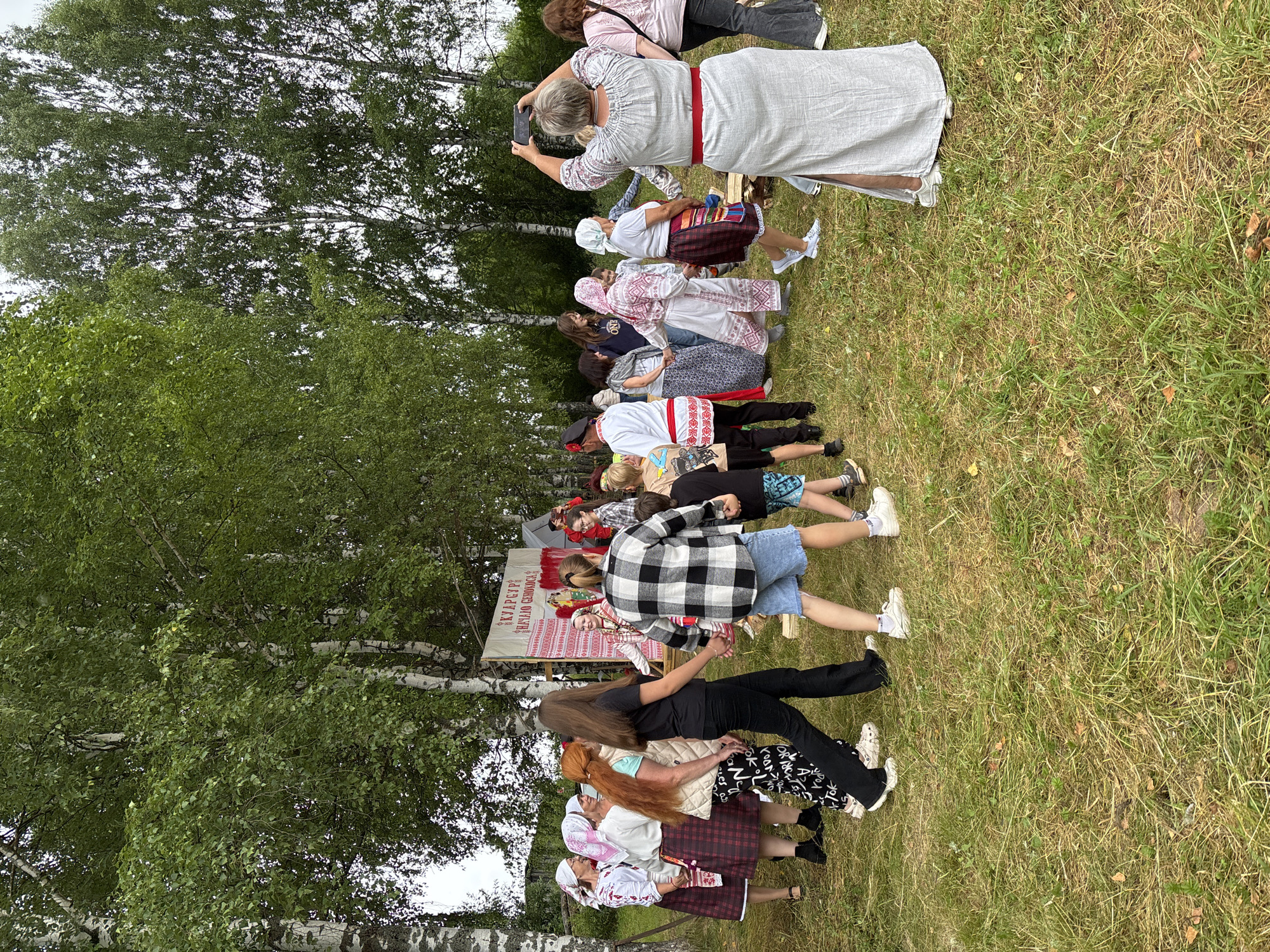 In a grassy clearing with birch trees in the background, a group of people are participating in a traditional folk event. Several women and girls are dressed in folk costumes, featuring embroidered shirts, skirts, and headscarves, some with intricate patterns and red accents. Some are holding hands and appear to be dancing or moving in a circle. One woman in a light-colored, long dress is capturing the event with her smartphone. A banner with Russian text is visible behind a section of the group. The ground is covered with dry grass, and there are some fallen leaves. The overall atmosphere suggests a cultural celebration or festival.