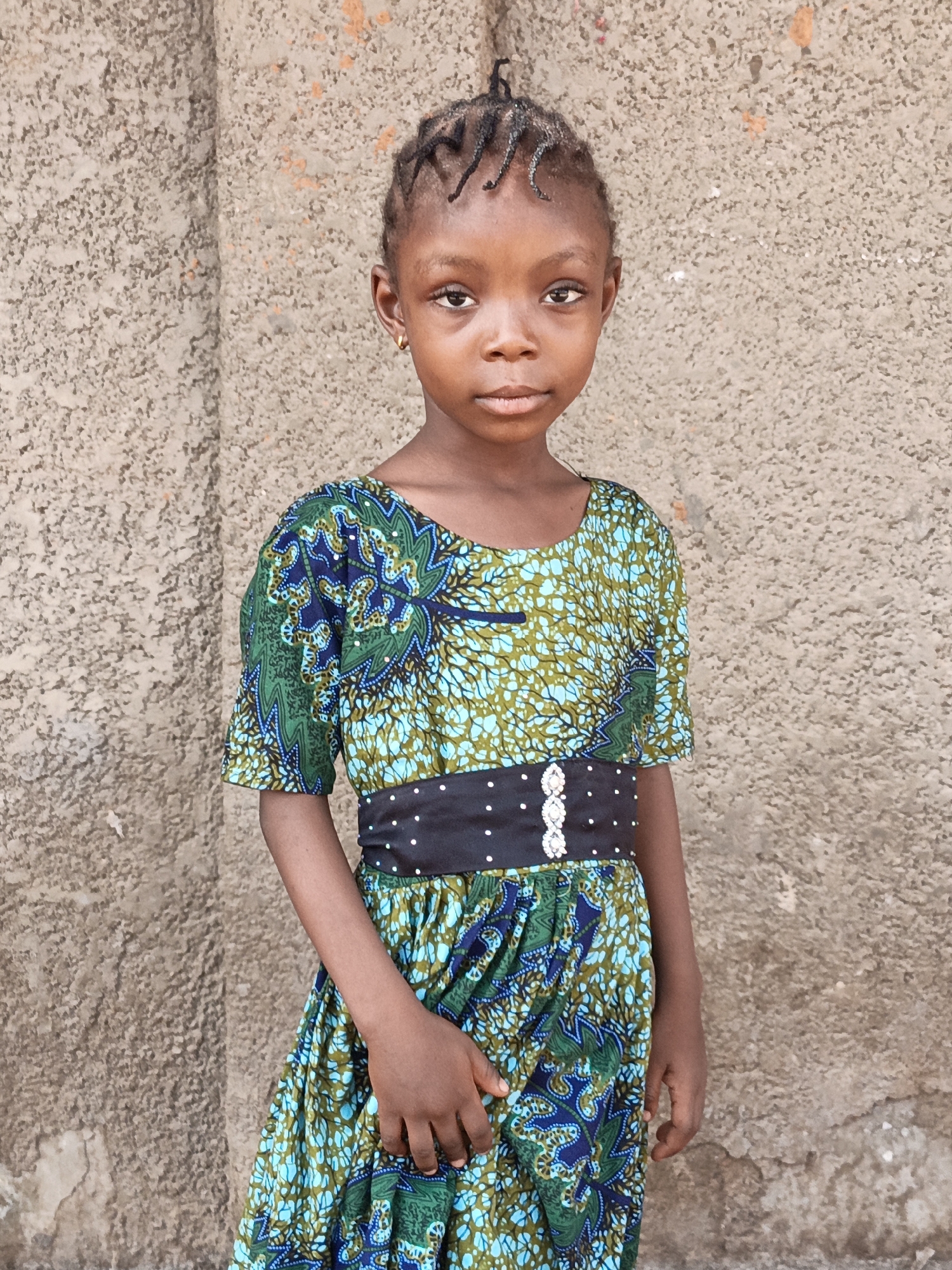 A young girl stands in front of a roughly textured, tan-colored wall. She has dark skin, short braided hair, and small gold hoop earrings. She wears a vibrant dress with a repeating pattern of green, blue, and light blue, cinched at the waist with a black belt dotted with small white embellishments. The location is Gawuna, Nigeria.