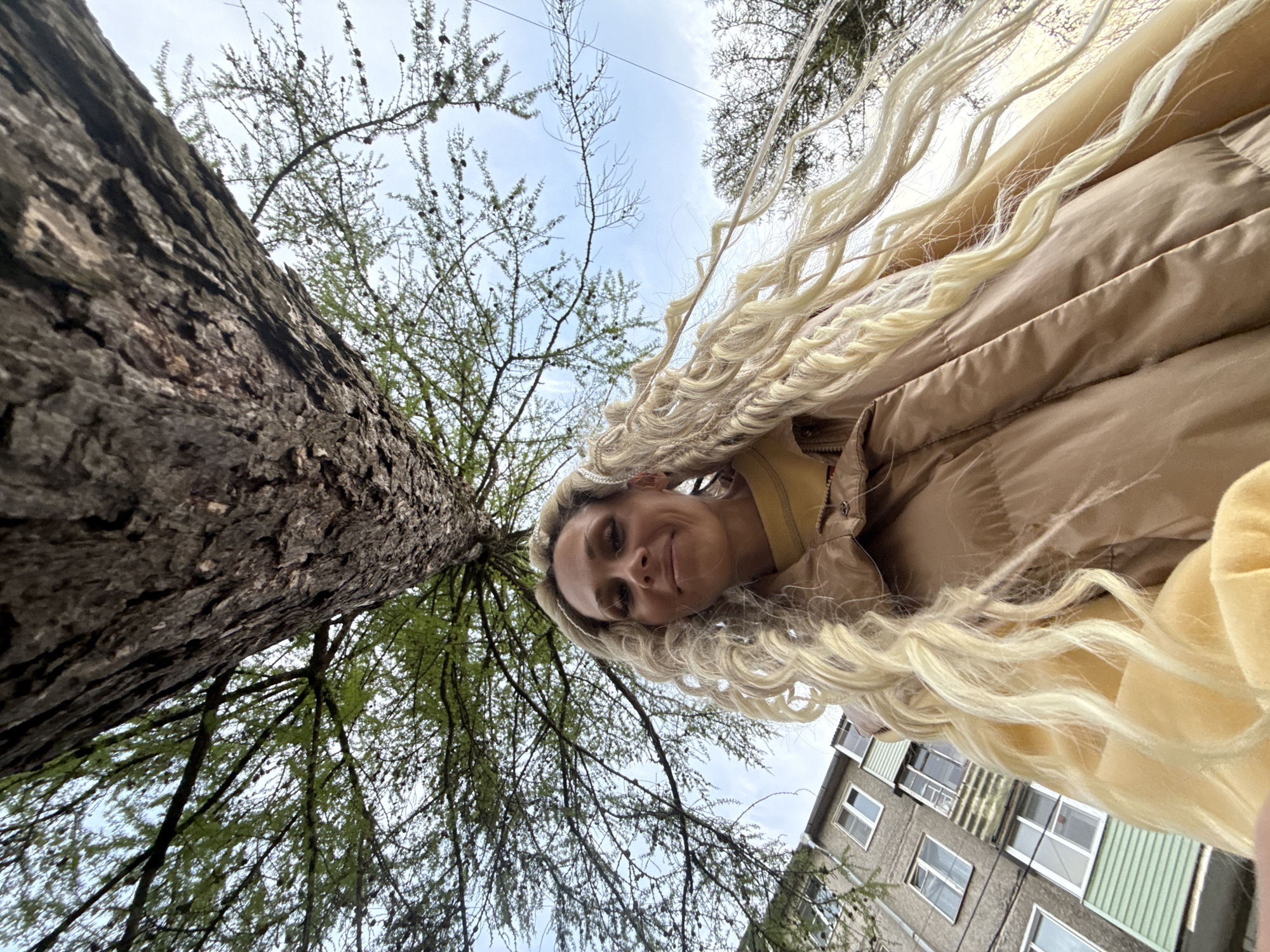 A low-angle outdoor shot shows a woman in a light brown puffer jacket and long, blonde, braided hair looking down at the camera with a slight smile. The angle suggests the photo was taken from a ground-level perspective. To the left, the rough bark of a large tree trunk fills a significant portion of the frame, with tree branches and sparse green leaves extending upwards towards the sky. A multi-story residential building with white-framed windows and green siding is visible in the lower right corner. The sky is overcast with a mix of blue and gray tones. Given the description, it's likely this photo was taken in or near Rusanovo, Russia.