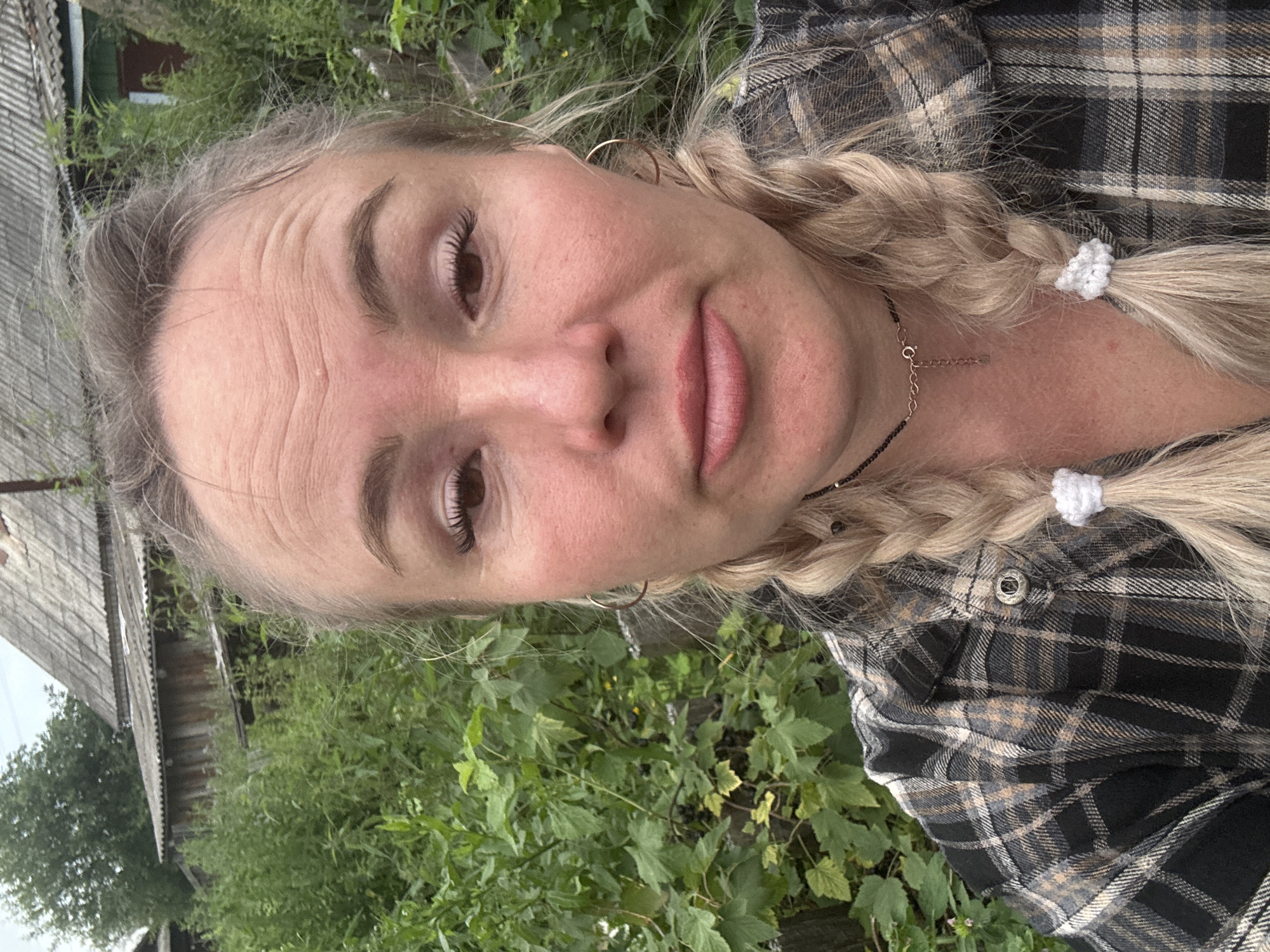 A young Caucasian woman with blonde hair in two braids and wearing a plaid shirt poses for a selfie outdoors. She is wearing hoop earrings and a choker necklace. The background features lush green foliage and a rustic wooden building, consistent with a rural setting in Nozhovka, Russia. The lighting suggests an overcast day.