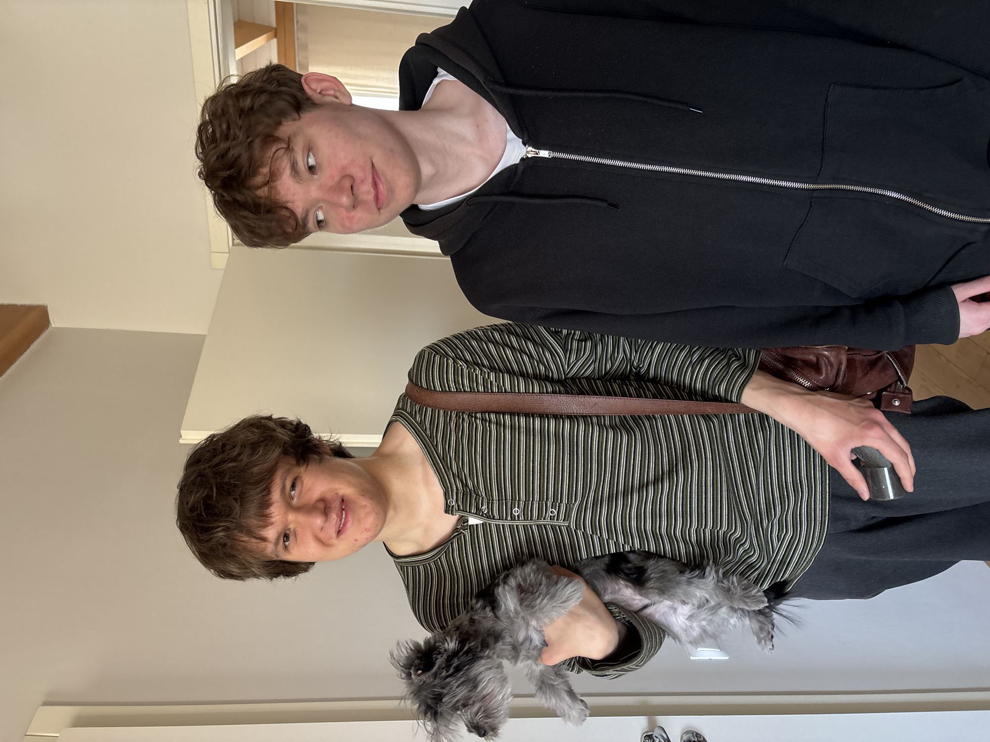 The image shows two young males and a small dog standing indoors, likely in a home setting in Chaikovsky, Russia. The room features plain, light-colored walls, suggesting a minimalist interior.
In the foreground, a young male with brown, medium-length hair and a slight smile is holding a small, gray, fluffy dog that resembles a Miniature Schnauzer. He is wearing a long-sleeved shirt with alternating vertical stripes in shades of green, black, and white, featuring a partial button-down placket. A brown leather shoulder bag is visible across his body, and he holds a small, silver cylindrical object in his right hand. The dog appears calm and well-groomed, with some areas of its fur appearing shaved.
Behind and slightly to the right of the first person stands another young male with short, curly brown hair. He is wearing a black zip-up hoodie over a white t-shirt, with the hoodie's zipper partially open. He looks towards the camera with a neutral, slightly curious expression.
The activity captured is a casual group portrait. The lighting is bright and even, indicative of daytime. There is no discernible text visible in the image. The overall mood is relaxed and informal.