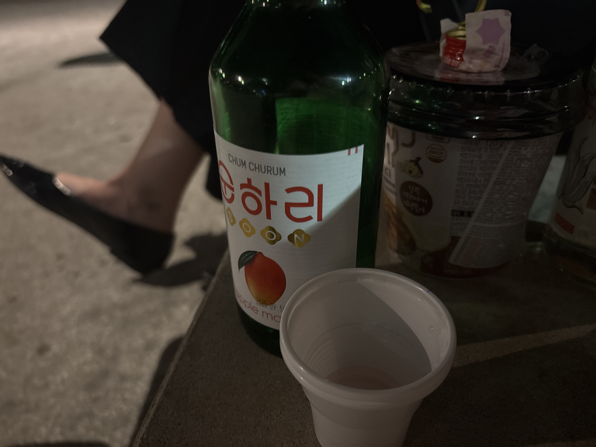The image depicts a casual outdoor setting with a bottle of Chum Churum soju, a white plastic cup with a small amount of liquid in it, and a food container. The soju bottle has a label indicating it is mango-flavored. A person's foot, wearing a black flat shoe, is visible in the background. The scene appears to be on a ledge or step, with dark, possibly outdoor lighting. There is no definitive information from the image to confirm the location is Milan, Italy.