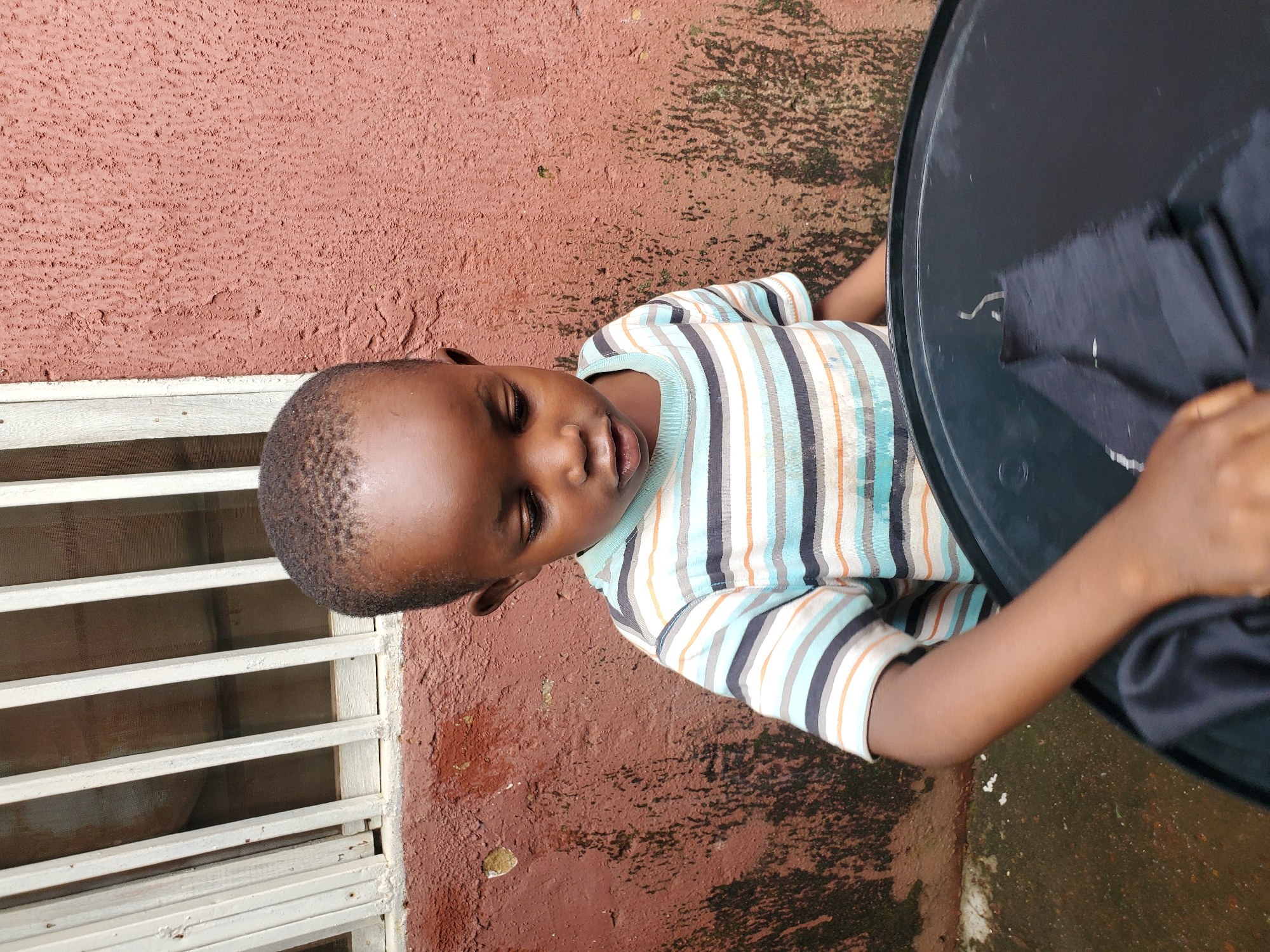 A young boy, with closed eyes and his mouth slightly open, is lying down, possibly resting. He is wearing a striped t-shirt with blue, white, orange, and black lines. His head is tilted to the right, and his right arm is resting on a large, black, circular object, which appears to be the surface of a table or a tray. A piece of dark fabric is draped over the object near his hand. The boy is positioned against a textured, reddish-brown wall, and to his left, there is a white metal security grill. The ground beneath him is a mixture of reddish-brown and darker, possibly damp, areas. The scene suggests an outdoor setting, potentially a patio or a yard, with the lighting indicating it is daytime. The overall mood seems relaxed and peaceful.