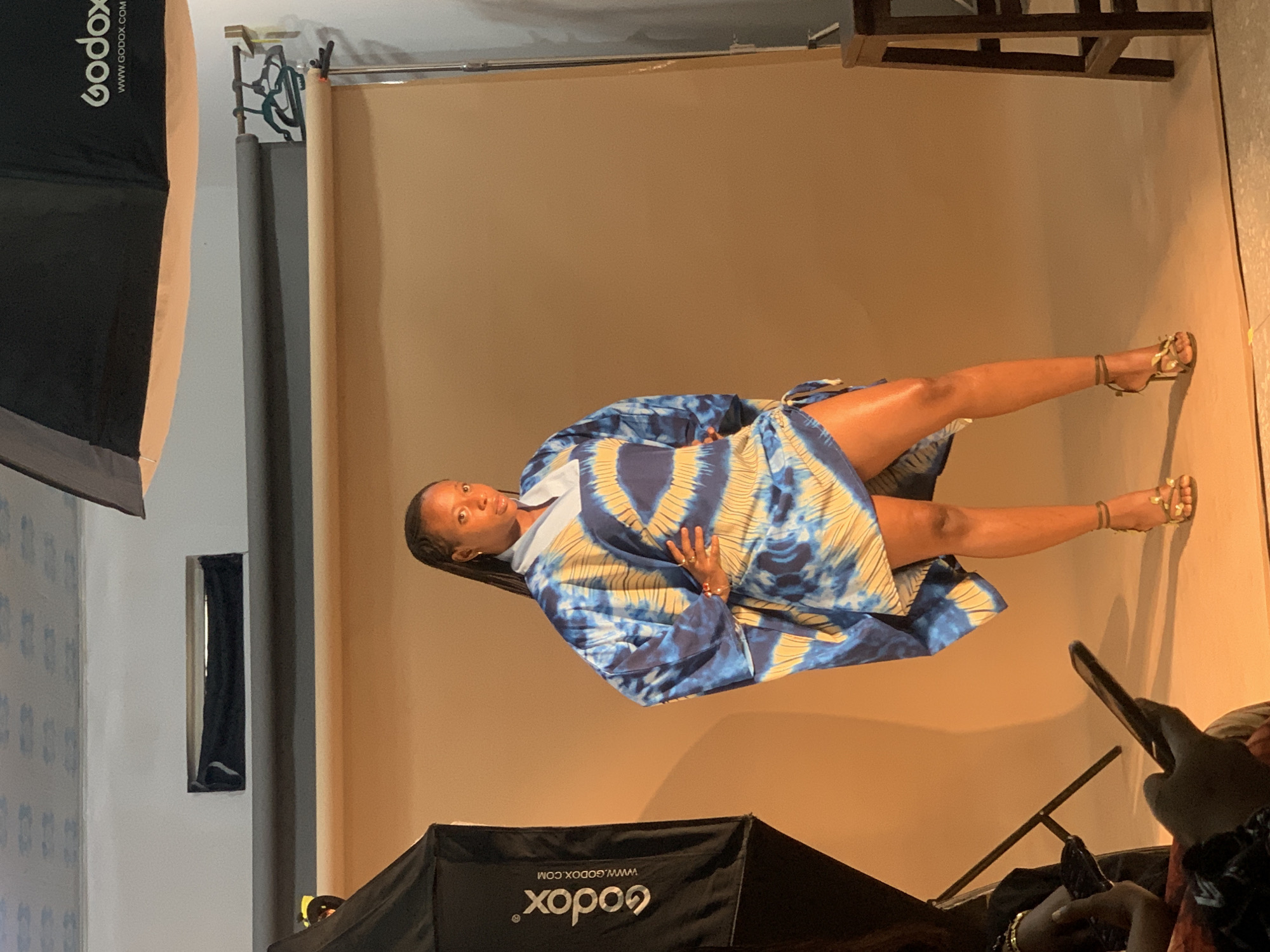 A woman poses in a studio in Gura Top, Nigeria. She wears a blue and yellow patterned dress and gold sandals. She stands in front of a light brown backdrop. A softbox is positioned to the left and in the foreground. A secondary light source and a ladder are visible in the background. An individual holds a cell phone in the foreground.