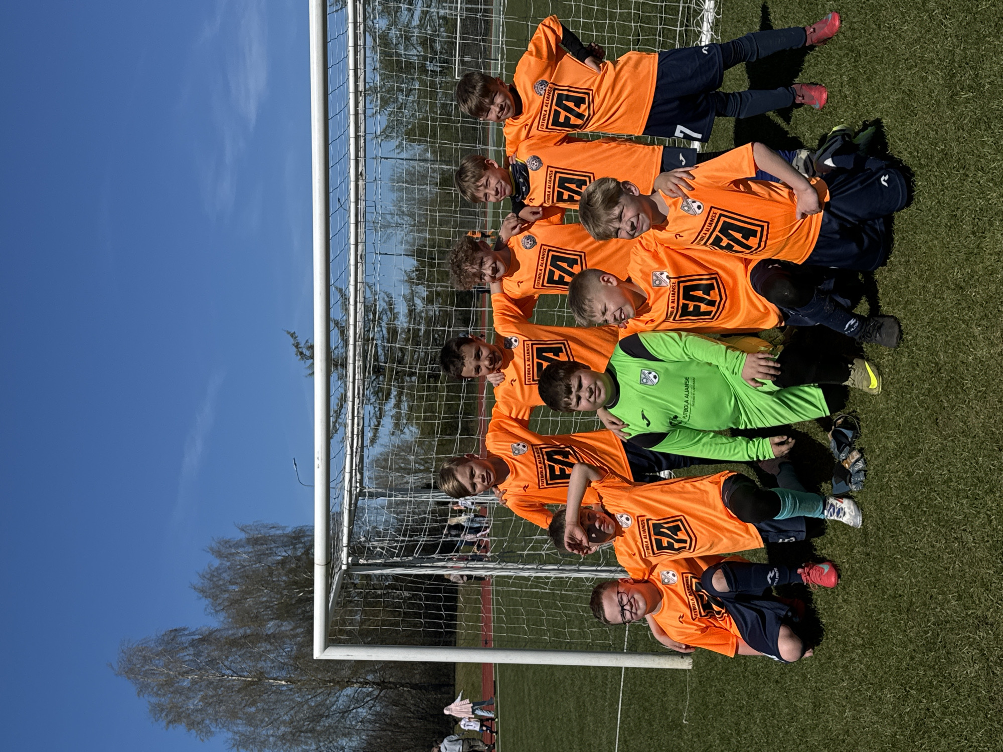 A youth soccer team is arranged in a portrait style lying against a soccer goal on a green sports field on a sunny day. The team consists of nine young boys. Eight of the boys are wearing orange soccer jerseys with "FUTBOLA ALIANSE" printed on them and blue shorts. One of the boys is wearing a green goalie jersey and black shorts. The soccer goal has a white frame and a white net. Blue sky is visible behind the goal. Bare tree branches are visible next to the goal, and additional people are in the background. The location is in Ragana, Latvia, as indicated by the jersey lettering.