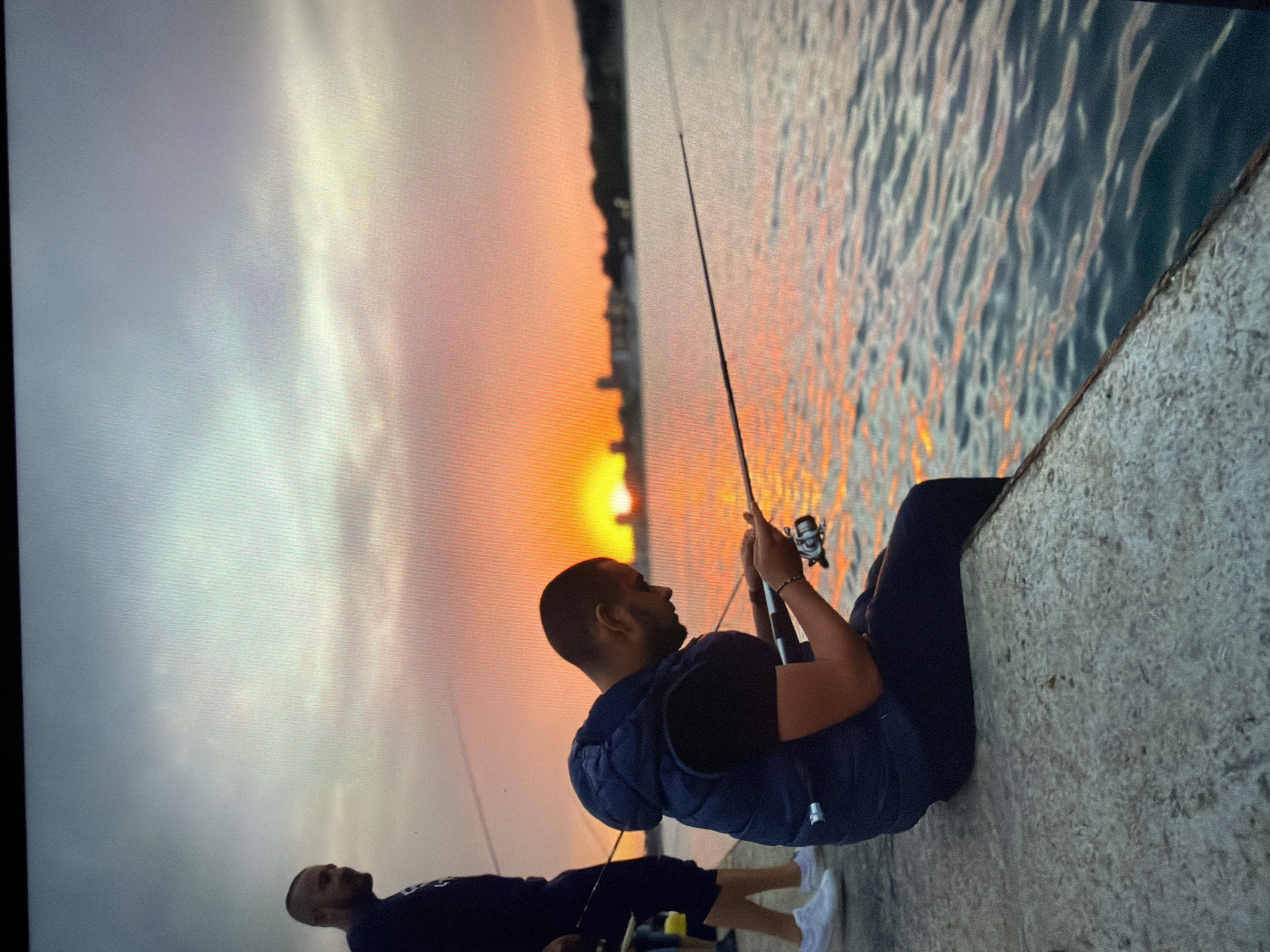 Two men are fishing at sunset in Burgas, Bulgaria. One man is seated on a concrete pier, holding a fishing rod and reel. He is wearing a dark blue vest and pants and white shoes. Another man is standing beside him, also holding a fishing rod. The sun is setting over the water, casting an orange glow on the scene. The silhouette of the city skyline is visible in the background. The water is reflecting the orange and yellow hues of the sunset.
