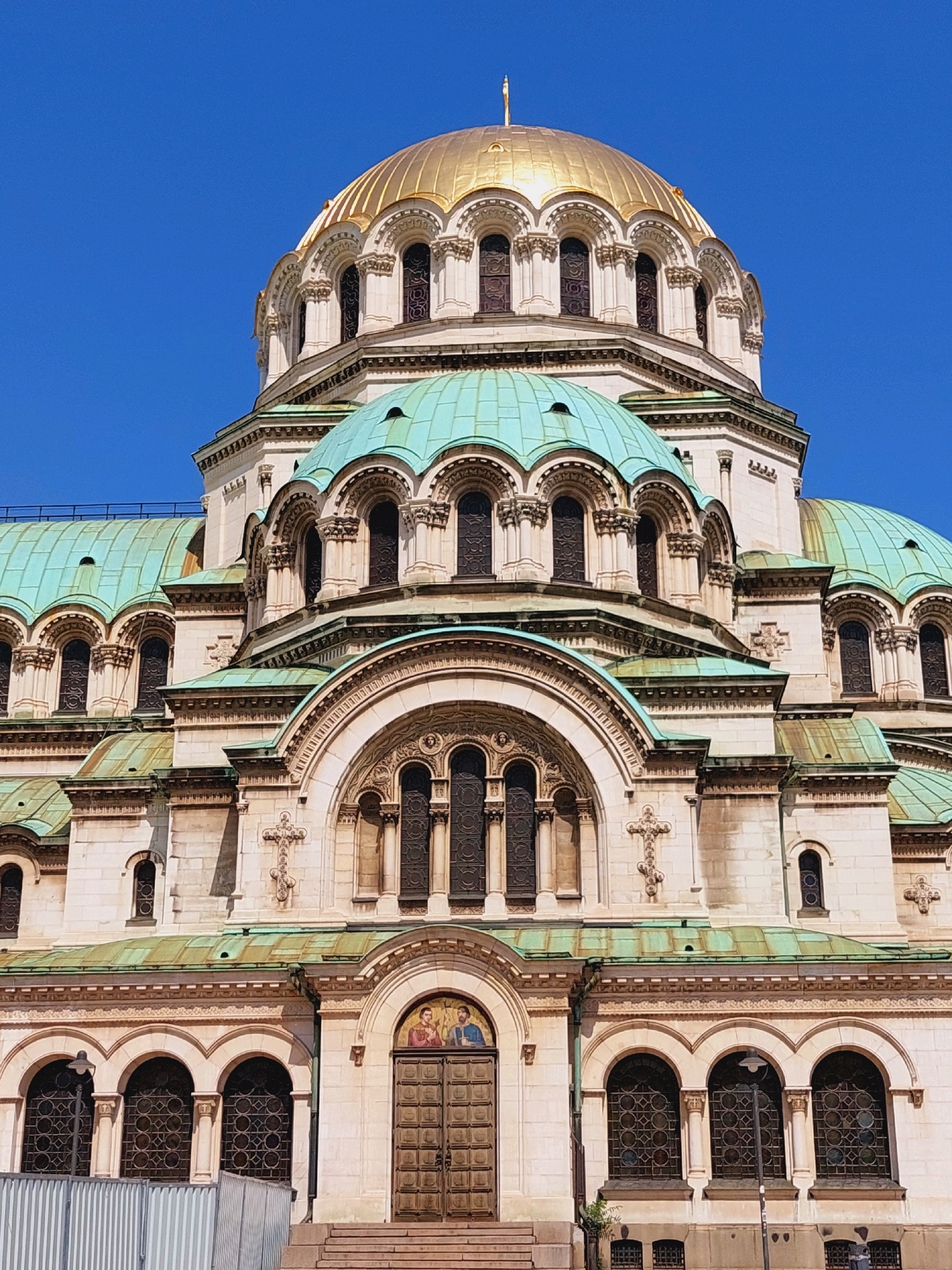This is a full-frame, eye-level, outdoor, vertical shot of the exterior of the Alexander Nevsky Cathedral, a prominent landmark in Sofia, Bulgaria. The cathedral, constructed primarily from light-colored stone, features multiple domes, with the central dome being large and gilded, topped with a cross. Smaller domes are covered in a verdigris-colored metal. The facade is adorned with intricate architectural details, including arched windows with stained glass, decorative carvings, and crosses. The sky above is a clear, bright blue. The lower portion of the image shows a large, ornate wooden double door, framed by arched windows. Steps lead up to the entrance. In the foreground, a metal fence can be seen on the left, and a lamppost stands near the windows on the right. No people are visible in the image.