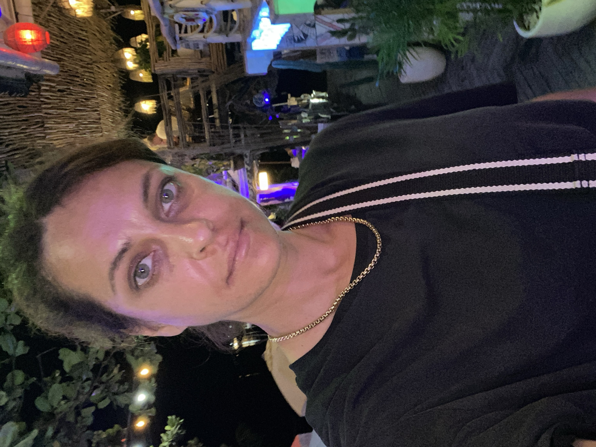 This is a close-up, vertically oriented, selfie-style photograph of a woman taken at night. She is wearing a black t-shirt with two white stripes across the chest and a gold chain necklace. Her hair is dark and pulled back, and she is looking towards the camera with a neutral expression. Her eyes are open and appear to be grey or blue. The lighting in the image is a mix of artificial lights from the background and ambient light, creating a pinkish hue on her skin.
The background is dimly lit and appears to be an outdoor setting with some thatched structures and hanging lanterns. There are also some green plants and string lights visible. The overall impression is that of a casual evening setting, possibly at a restaurant or bar. The location context provided is Nusa Penida, Indonesia. While the image itself does not contain any visible text, the setting suggests a tropical or semi-tropical environment, and the time of day is clearly night due to the darkness and the prevalence of artificial lighting.
