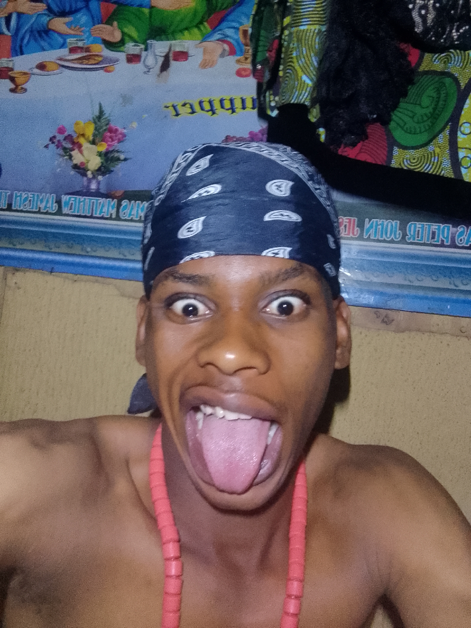 A dark-skinned, bare-chested young man with a black bandana and a red beaded necklace is pulling a face with his tongue sticking out. The background includes a wall with a light brown texture and a poster depicting the Last Supper. The location is Nkpor, Nigeria.