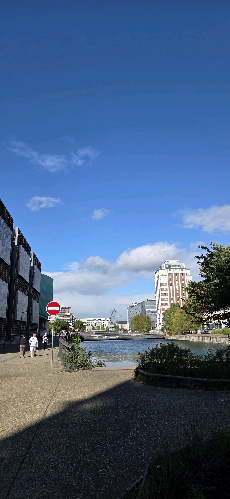 A vertical, eye-level, medium-wide shot captures a sunlit, urban waterfront scene in Strasbourg, France. The image is dominated by a clear blue sky with scattered white clouds. In the foreground, a grey, speckled pedestrian pathway curves from the left towards the right, casting a sharp shadow that covers the lower right quadrant. On the left side, the modern facade of a multi-story building with metallic siding and rectangular panels is visible. Further along the pathway, three individuals are walking away from the camera, two men in casual attire and a woman in a patterned top and white shorts. A red and white circular "no entry" sign stands on a pole to the right of the pathway.
The pathway leads to the edge of a calm body of water, likely a canal or river, with a low metal railing separating it from the walkway. On the opposite side of the water, a cityscape unfolds. A distinctive white and red-striped building with numerous windows stands prominently. Adjacent to it is a grey, modern building. In the background, a large industrial crane is visible, suggesting a port or industrial area. Trees with green foliage are scattered throughout the scene, particularly around the buildings on the far bank. The overall impression is one of a bright, clear day in a European city.