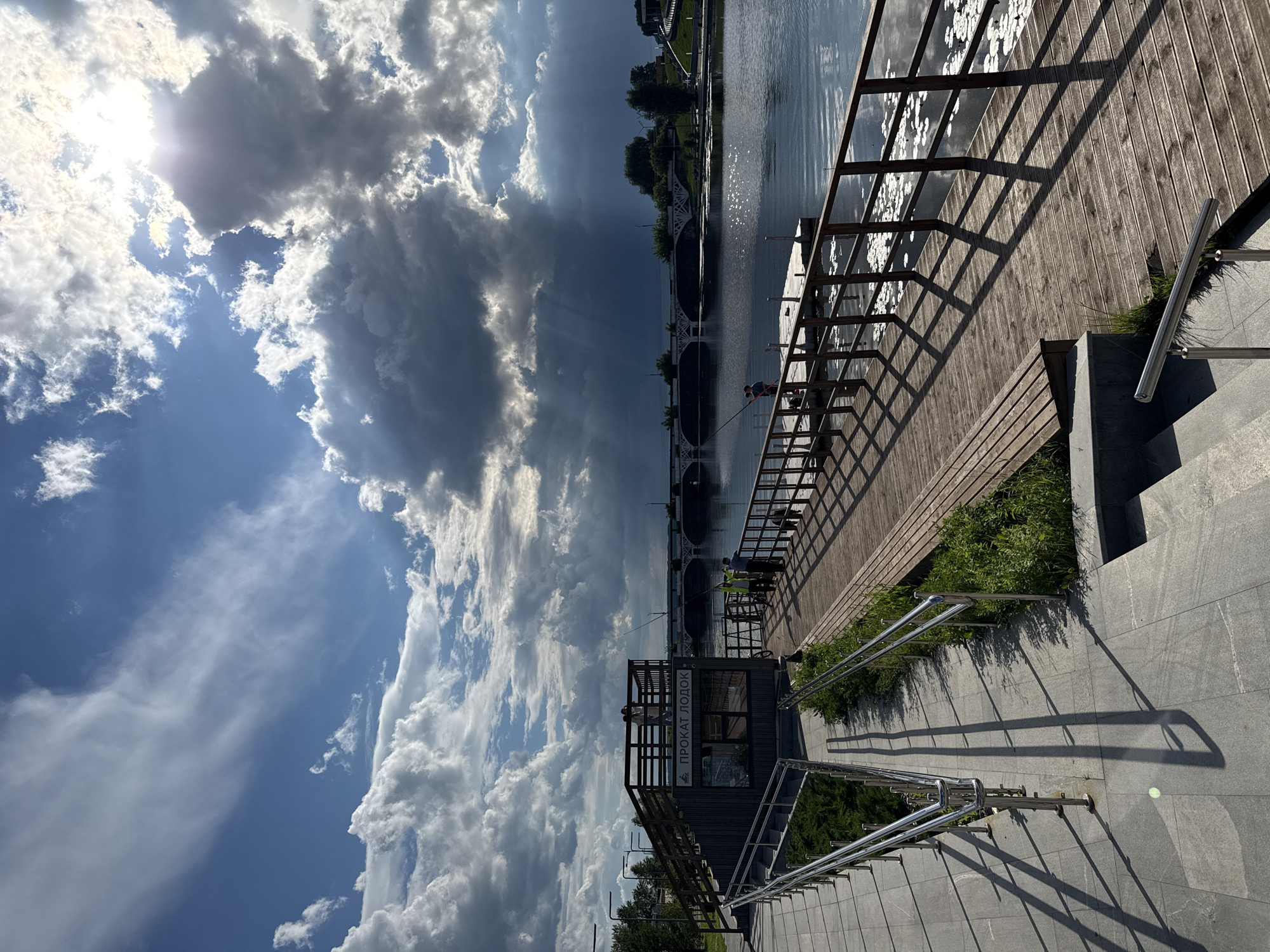 In Votkinsk, Russia, a wide wooden pier extends into a calm body of water under a dramatic sky filled with a mix of bright sunlight and dark, heavy clouds. The sun peeks through the clouds, casting rays of light onto the water and pier. On the pier, several individuals are engaged in fishing, their silhouettes visible against the water. To the left of the pier, a modern walkway with metal railings leads down to the water's edge. Adjacent to the walkway, a building with a sign that reads "ПРОКАТ ЛОДОК" (Boat Rental) is situated. Across the water, a bridge with multiple arches spans the distance, and beyond it, the Votkinsk dam is visible in the background. The environment appears to be a waterfront recreational area on a partly cloudy day.