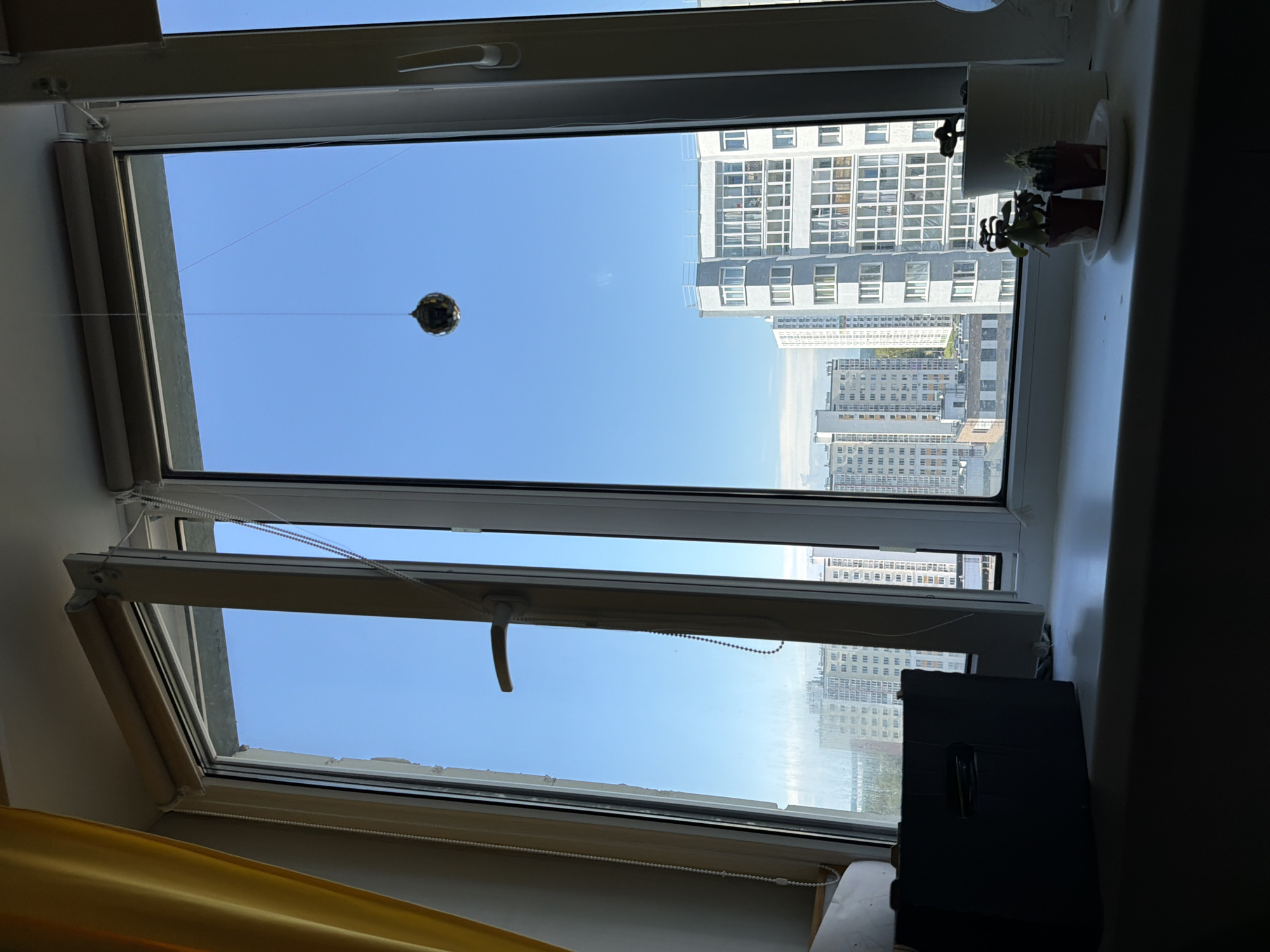 A medium shot shows a view from a window looking out over an urban landscape in Ufa, Russia. The window itself is a casement-style window with a pane that is partially opened. A yellow curtain is visible to the left of the frame. In the foreground, on the windowsill, are a dark rectangular object, two small potted plants, and a white paper towel dispenser. A small, reflective, ball-shaped ornament hangs from a string in front of the window, suspended against a clear blue sky. In the background, a series of multi-story apartment buildings are visible, characteristic of a Russian urban environment. The buildings are predominantly light-colored with many windows. The overall scene conveys a sense of everyday life within a residential area.