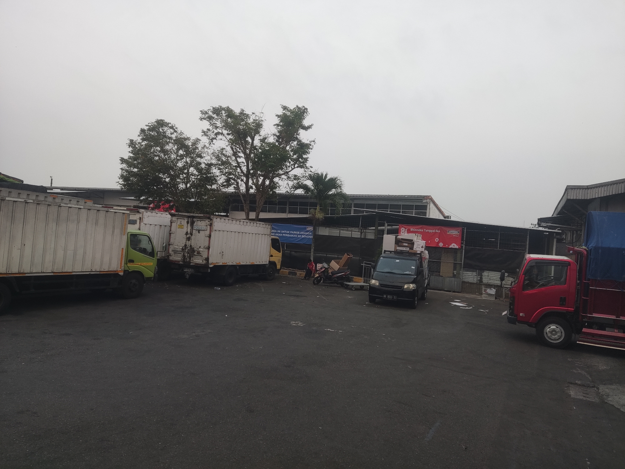In Trihanggo, Indonesia, an overcast day features an industrial area with multiple trucks parked on an asphalt lot. To the left, a white box truck with a light green cab is positioned. Adjacent to it, another white box truck with a yellow cab is parked. Further to the right, a black van with an Indonesian license plate "B 8533 EU" is present. A red truck with a blue tarp covering its cargo bed is seen on the far right. In the background, a long building with a gray roof and a row of windows is visible. Several trees, including palm trees, are scattered throughout the scene. A banner with text in Indonesian is hung on the building. The ground is wet in several patches, suggesting recent rain. There are a few scattered objects, including some cardboard boxes and a motorcycle.