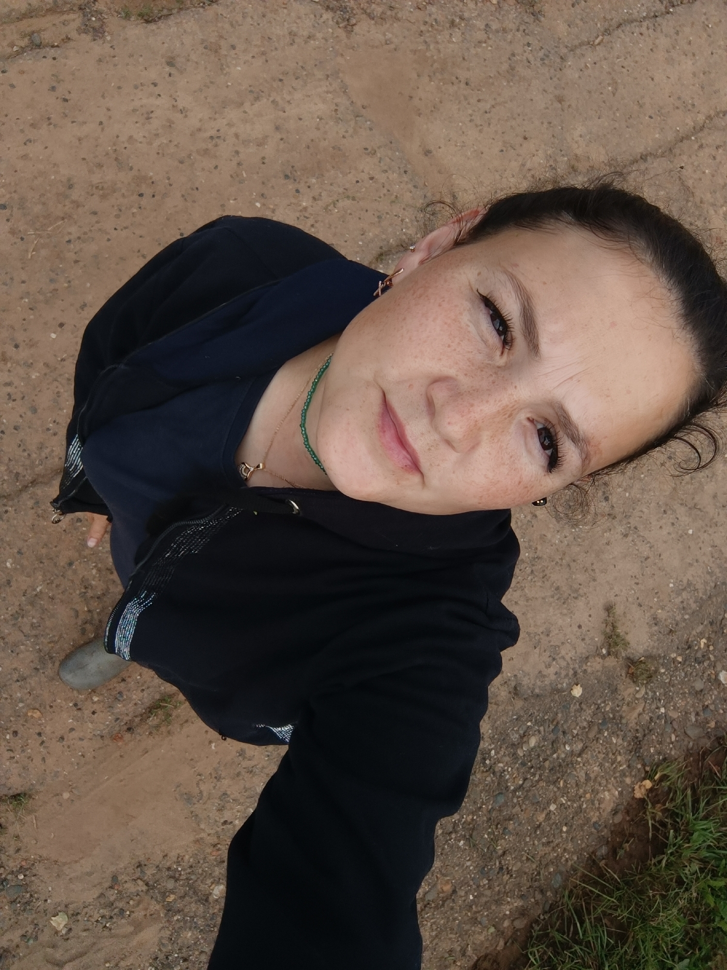 The image is a high-angle, medium shot of a woman outdoors, looking upwards at the camera. She has dark hair pulled back and prominent freckles on her face. She is wearing a dark blue t-shirt, a black zippered hoodie, and light grey footwear. A thin, green beaded necklace is visible around her neck, with a gold pendant. The ground beneath her is composed of dirt and small stones, with some grass visible on the right side of the frame. The lighting suggests it is daytime. No specific landmarks of Karsha, Russia are identifiable in this image.