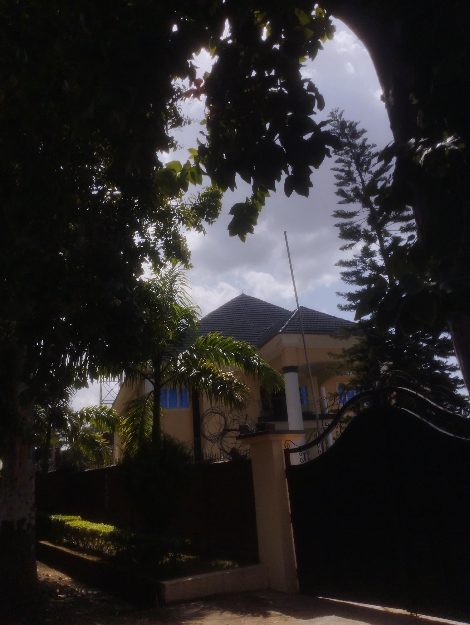 The image depicts a large, modern house framed by dark foliage. The house has a tiled roof and cream-colored walls with bright blue windows. A black, ornate gate stands in the foreground, partially obscuring the view of the property. There is a well-maintained hedge in front of a dark wall.
The scene appears to be set in a tropical or subtropical region, suggested by the presence of palm trees. The sun is partially obscured by clouds, indicating it is daytime, likely mid-morning or afternoon. The sky is a mix of bright clouds and darker, possibly stormy, clouds. The overall impression is one of a well-to-do residence in a verdant setting. No people or specific activities are visible. There is no visible text in the image.