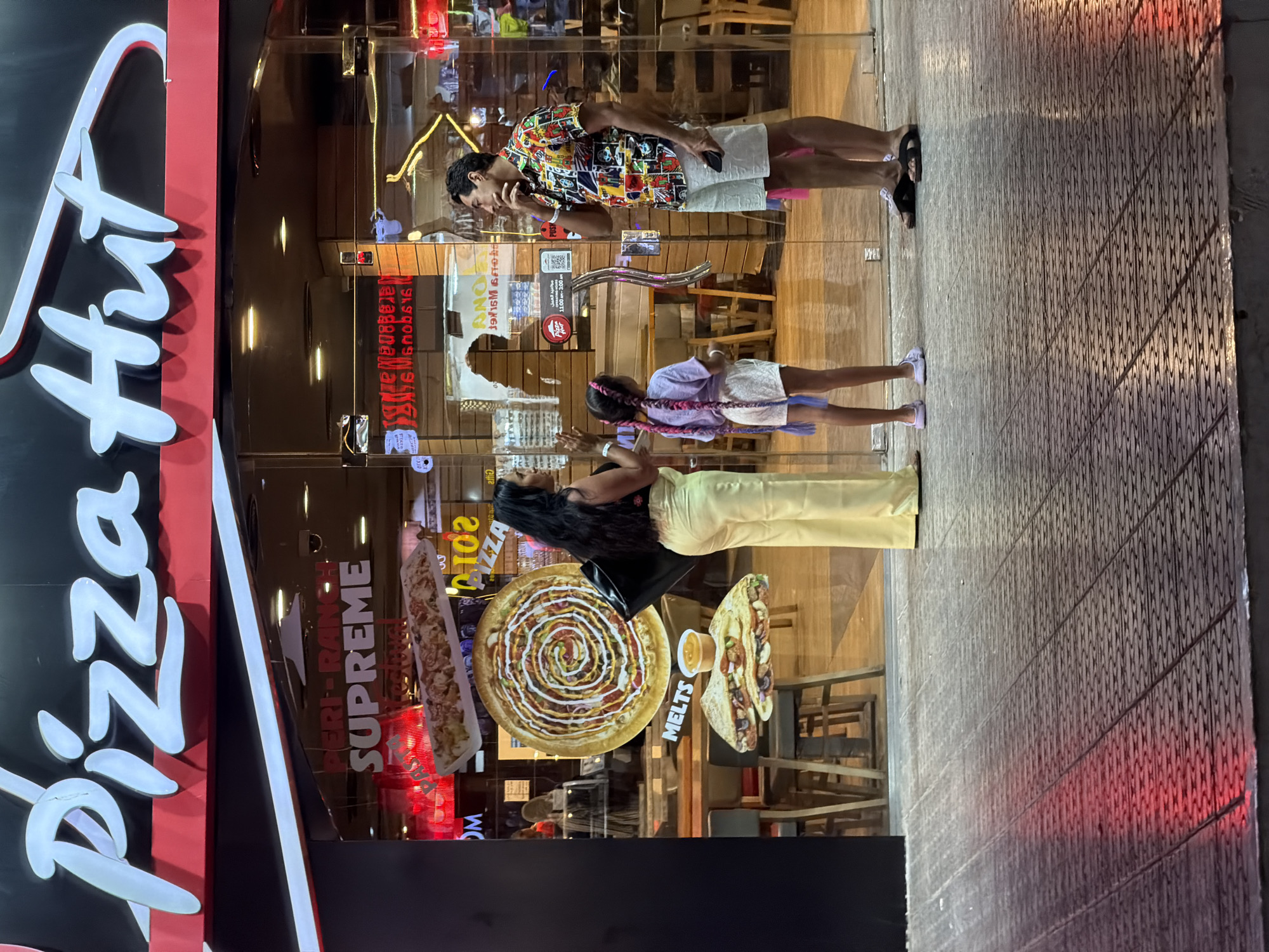 The image captures the exterior of a Pizza Hut restaurant in Sharm El Sheikh, Egypt. The prominent red and white Pizza Hut logo is visible above the entrance. Inside, through the glass doors, a family group is visible. A woman in light yellow pants stands facing away from the camera, holding a black bag. Beside her, a young girl with long, braided hair stands facing the entrance. A man wearing a colorful patterned shirt and white shorts stands near the door, looking thoughtfully to the side and holding a smartphone. Several promotional images of pizzas and melts are displayed on the glass, including a large circular pizza graphic with a spiral design and a depiction of a folded sandwich. The foreground shows a patterned tiled floor with red and yellow lighting reflecting on its surface. The overall scene suggests a casual dining atmosphere outside a popular fast-food chain.