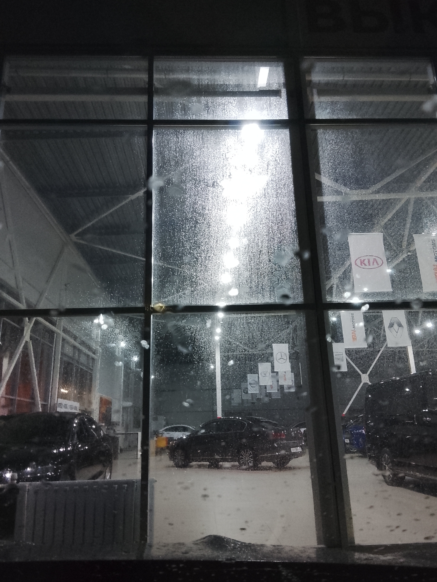 The image shows the exterior of a car dealership, likely located in Obninsk, Russia, viewed through a rain-covered window. The dealership is visible through the glass panels. Inside the dealership, several cars are parked. Banners with logos of car brands, including KIA, Mercedes-Benz, and Renault, are visible. The environment suggests it is nighttime or a dimly lit evening with rain falling.