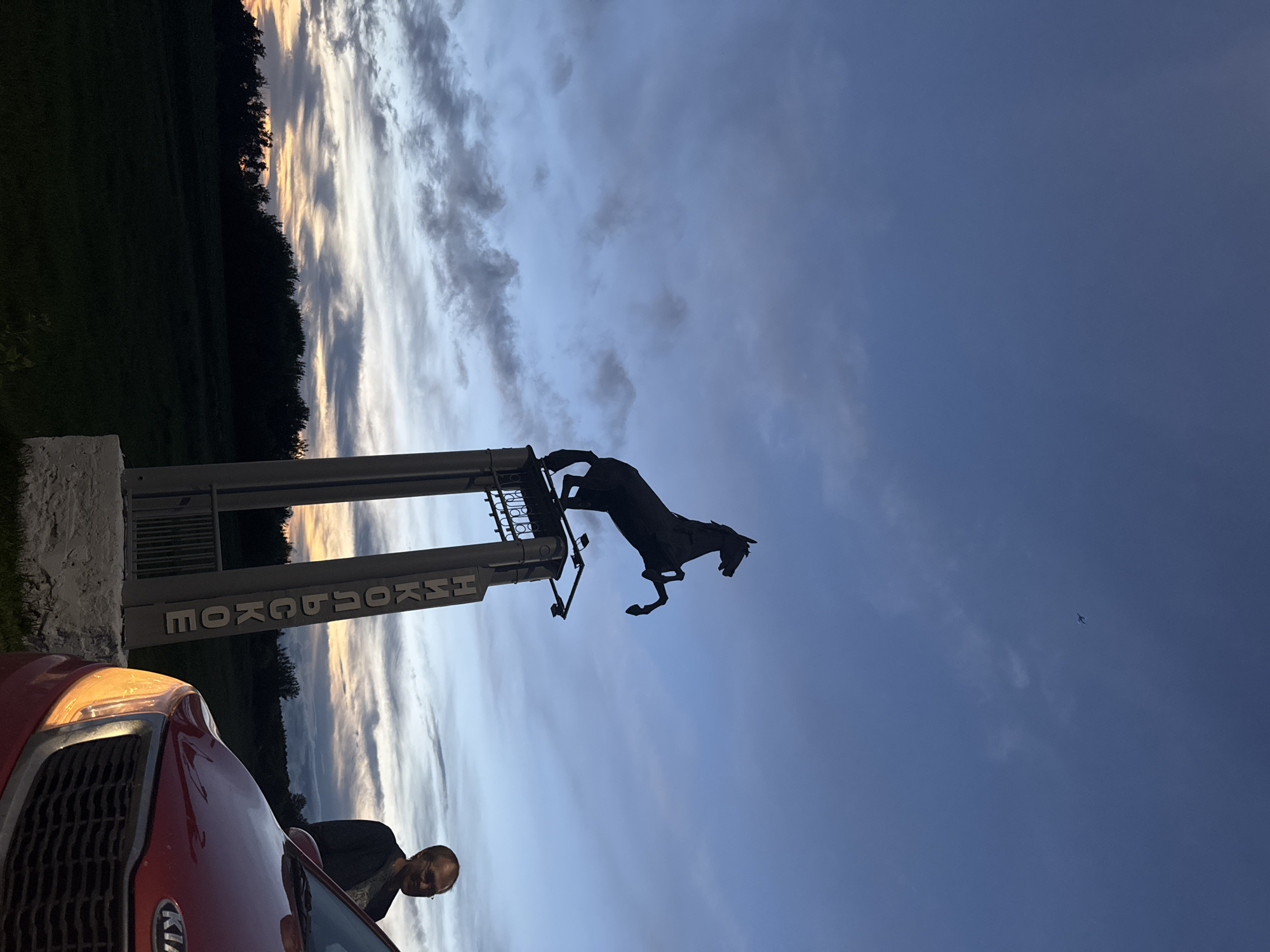 A silhouette of a horse statue stands on a tall pedestal against a dusky sky in the city of Nikolskoye, Russia. The statue depicts a rearing horse. Below the horse, on the pedestal, is a sign with Russian text that reads "НИКОЛЬСКОЕ" vertically. To the right of the pedestal, a man with glasses is partially visible, leaning against a red Kia car. The car's headlights are illuminated, casting a warm glow. The background shows a dark silhouette of trees and a field under a sky with streaks of clouds and remnants of sunset colors. The overall scene is set outdoors at dusk.