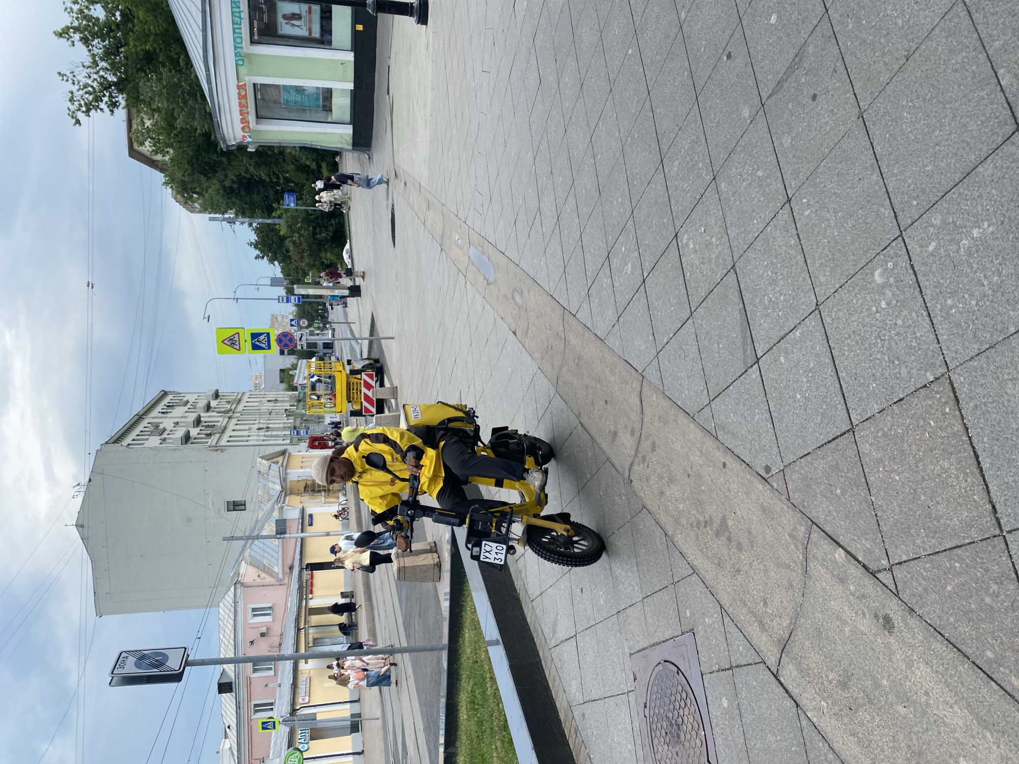 A person wearing a yellow jacket and a beige cap is riding a yellow electric scooter on a paved pedestrian walkway in Moscow, Russia. The scooter has a black box on the back with yellow text and a license plate with the alphanumeric characters YX7 310. The person is looking forward. In the background, there are buildings, trees, and other pedestrians. There are also traffic signs and power lines visible. The sky is overcast. The walkway is made of grey paving stones.