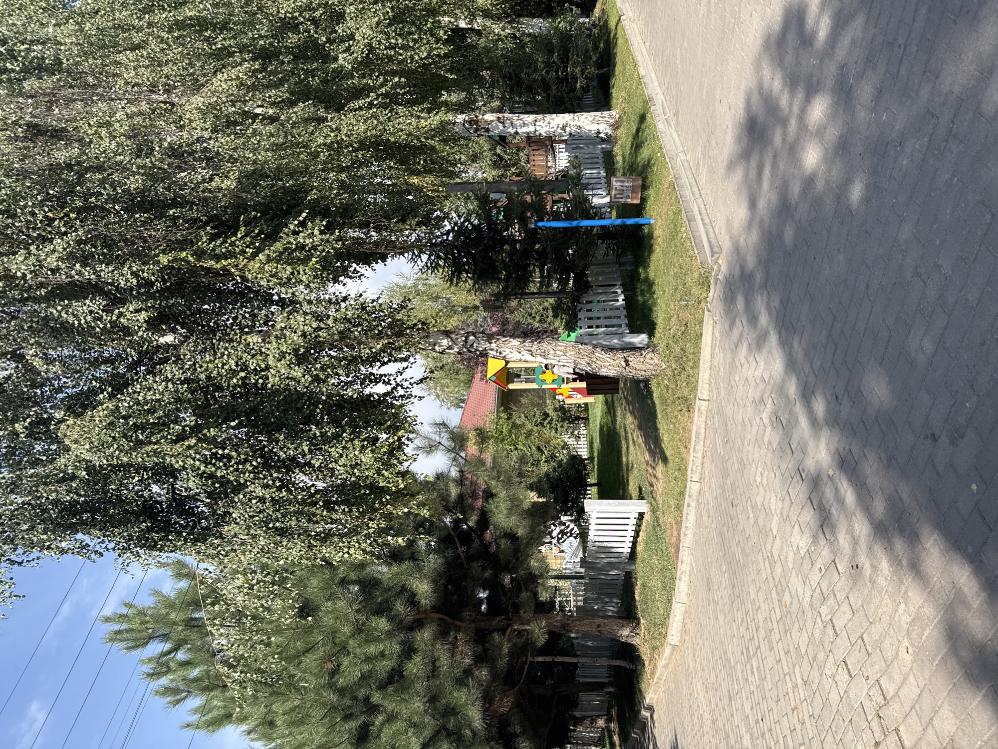 The image depicts a public outdoor area with a paved walkway in the foreground and a grassy area beyond, separated by a low curb. A white picket fence runs along the edge of the grassy area. In the background, there is a playground with colorful structures and a red-roofed building. Several trees, including pine and birch, are scattered throughout the scene, casting shadows on the ground. Power lines are visible in the upper left portion of the image against a blue sky with some clouds. A blue pole and a small sign with Cyrillic text are also present in the grassy area. The overall scene appears to be a park or recreational area.