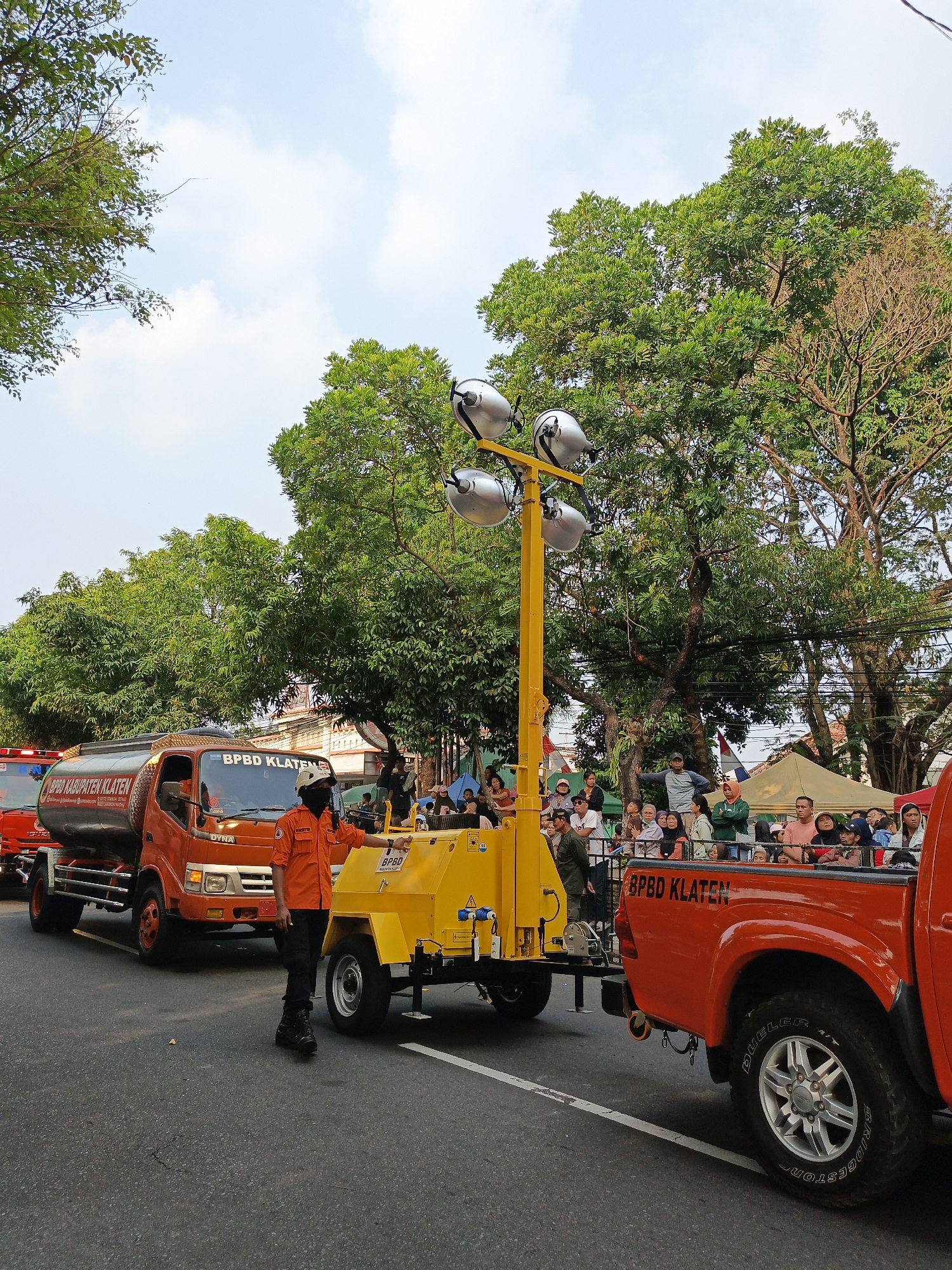The image depicts a daytime scene on a paved road in Gayamprit, Indonesia, likely during a parade or event. In the foreground, a person in an orange uniform, possibly a first responder, stands beside a yellow trailer with a large light tower mounted on it. The trailer is connected to an orange pickup truck with "BPBD KLATEN" written on the tailgate. To the left of the pickup truck is an orange tanker truck, also bearing "BPBD KABUPATEN KLATEN" and "BPBD KLATEN" on its side. The truck appears to be a water or fuel tanker.

A crowd of spectators is visible in the background, standing behind a fence or barrier, observing the procession. They are dressed in casual attire, and some are holding umbrellas. Lush green trees line the street, and the sky is visible with scattered clouds. The overall impression is of a community event with emergency services participating, set against a backdrop of urban foliage and a clear sky. The atmosphere appears festive and orderly.