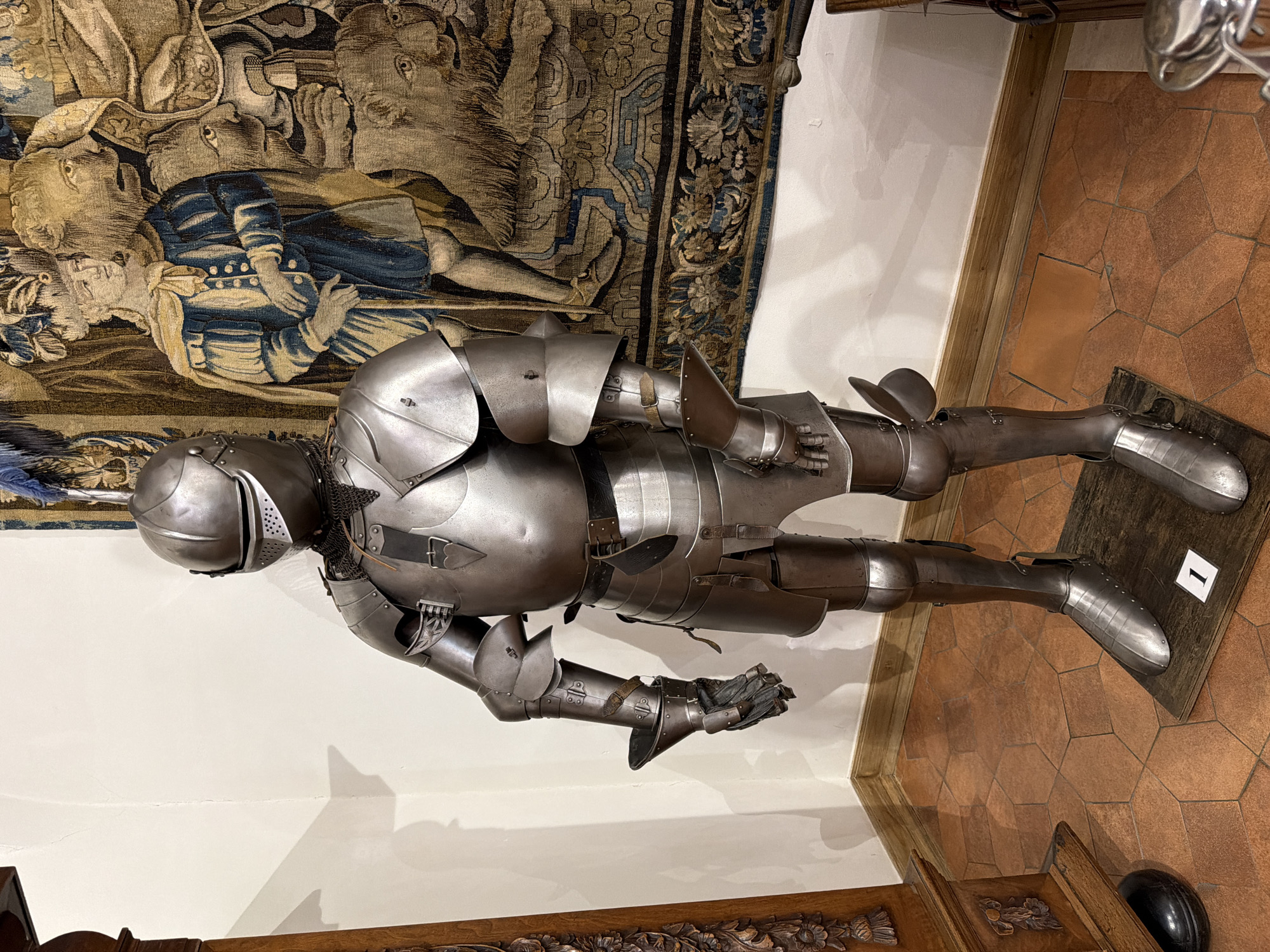 This media file shows a full suit of polished metal armor, likely a knight's armor from a historical period, displayed vertically. The armor covers the entire body, from the helmet to the sabatons (foot armor). It is detailed, with articulated joints and protective plates. The armor appears to be in excellent condition, reflecting light from an unseen source.
Behind the armor, a tapestry hangs on a wall. The tapestry depicts a scene with figures, possibly historical or mythical, rendered in muted colors of blue, brown, and beige. The style of the tapestry suggests it is also an antique.
The armor is positioned on a dark wooden base, which rests on a floor covered in hexagonal terracotta tiles. The lighting in the scene is neutral, suggesting an indoor display, perhaps in a museum or historical exhibition. There are no discernible people actively interacting with the armor, implying it is a static display.
A small white card with a black border and the numeral "1" is visible on the wooden base near the armor's right foot. This card likely serves as an exhibit label or number. The overall impression is one of historical preservation and presentation, highlighting the craftsmanship of both the armor and the tapestry. The scene is static and focused on the objects themselves.